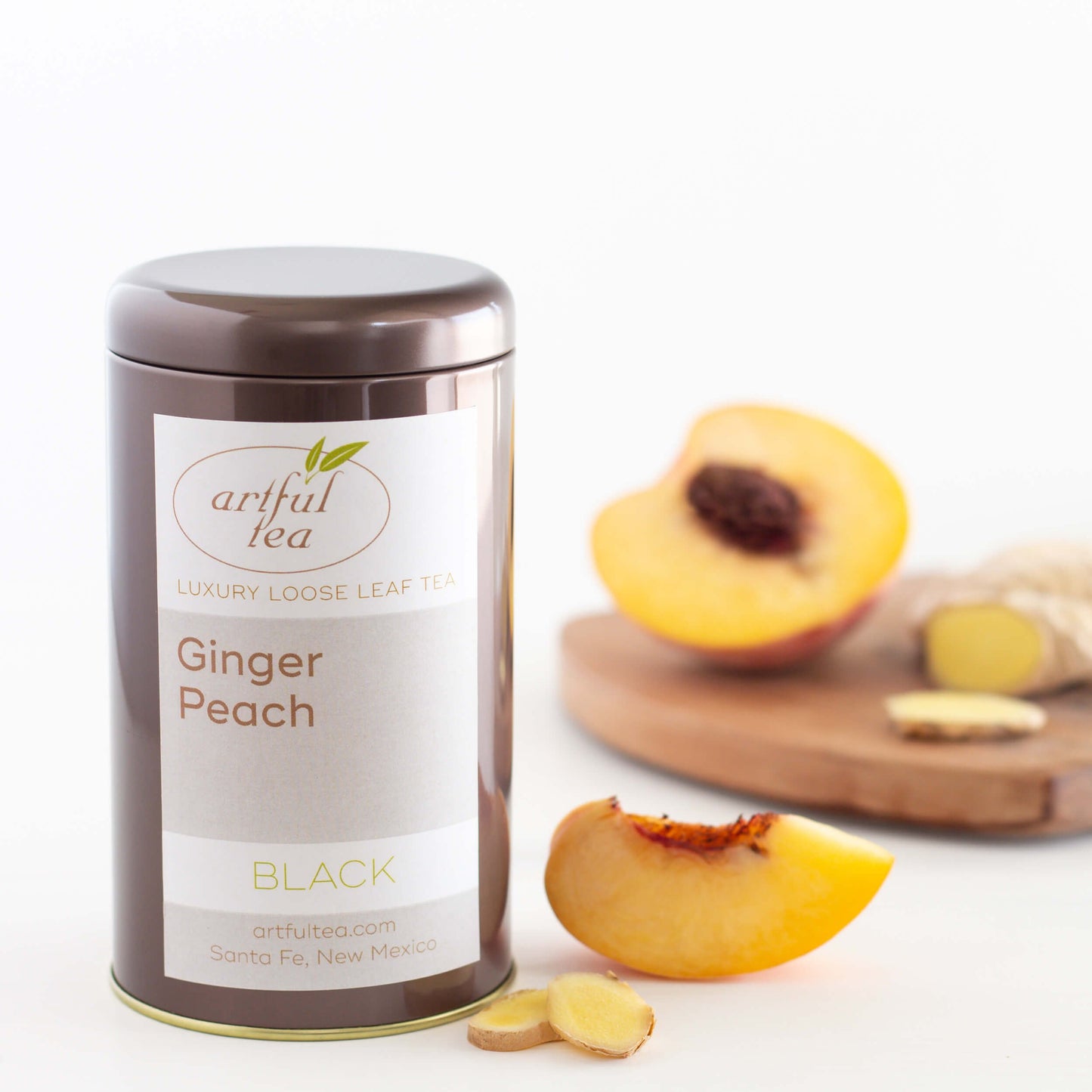 Ginger Peach Black Tea shown packaged in a brown tin next to a wedge of fresh peach and two slices of fresh ginger. A peach half and more ginger are displayed on a wooded board in the background