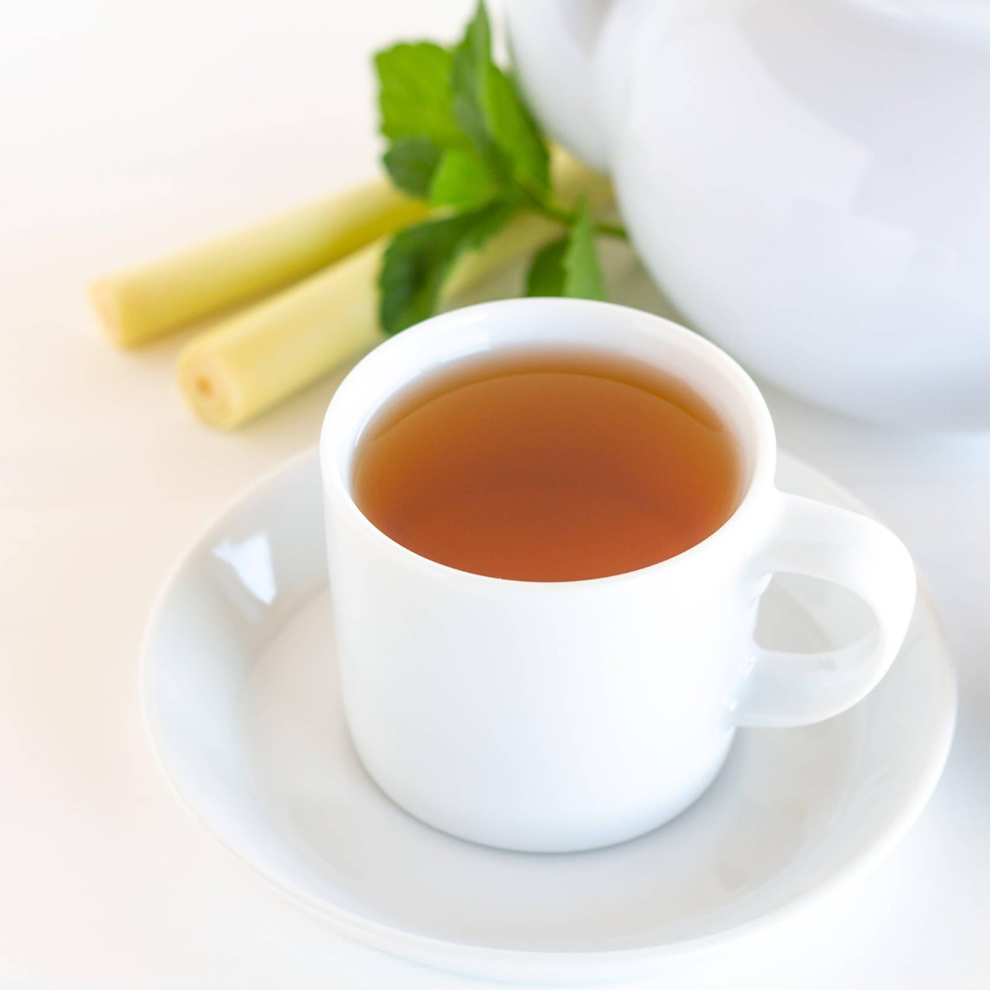 Lemon Mint Ménage Organic Black Tea shown as brewed tea in a white teacup and saucer, with a white teapot, lemongrass and mint leaves in the background