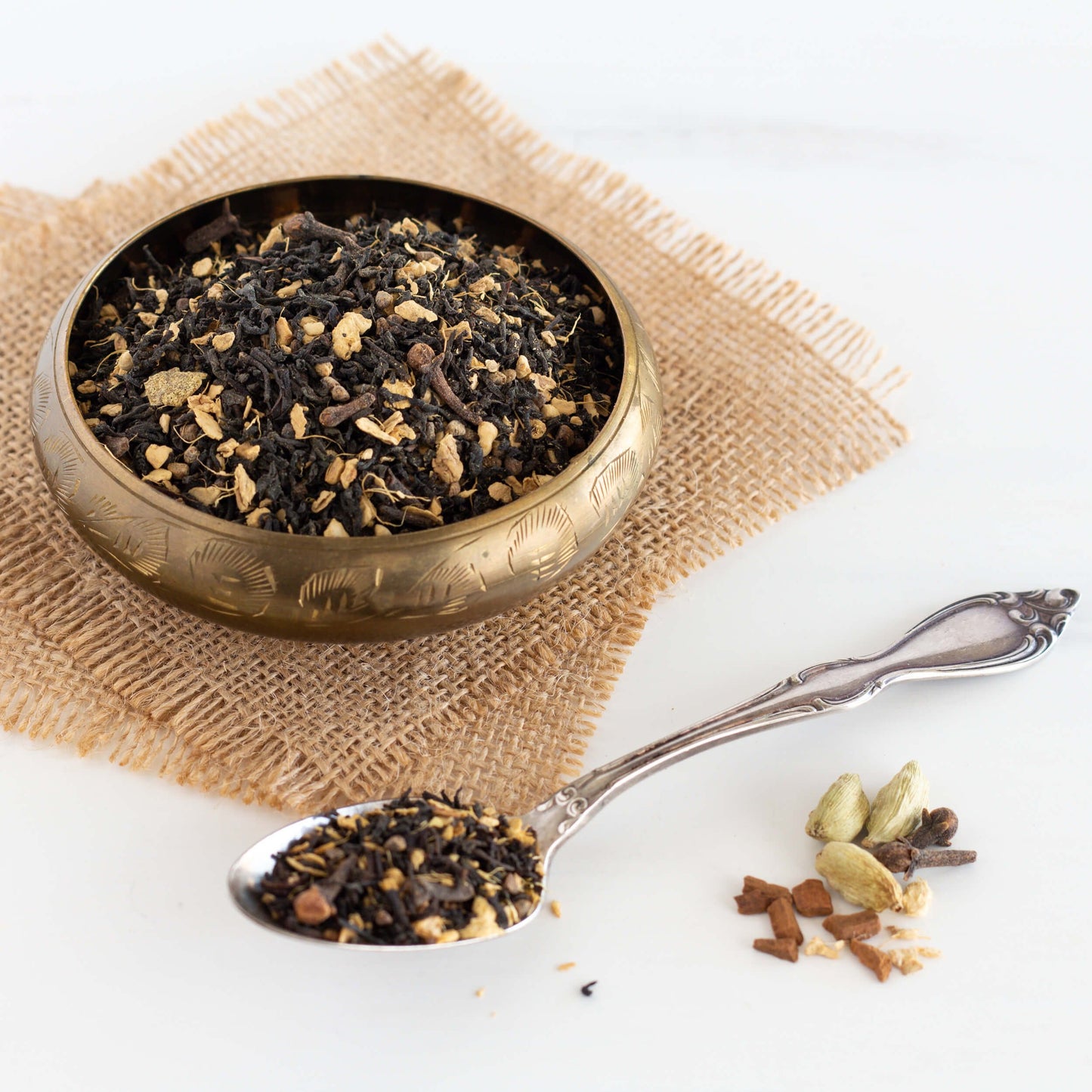Masala Chai Black Tea shown as loose leaf tea displayed in a small brass bowl on two squares of burlap. A silver spoon with tea leaves in it is in the foreground