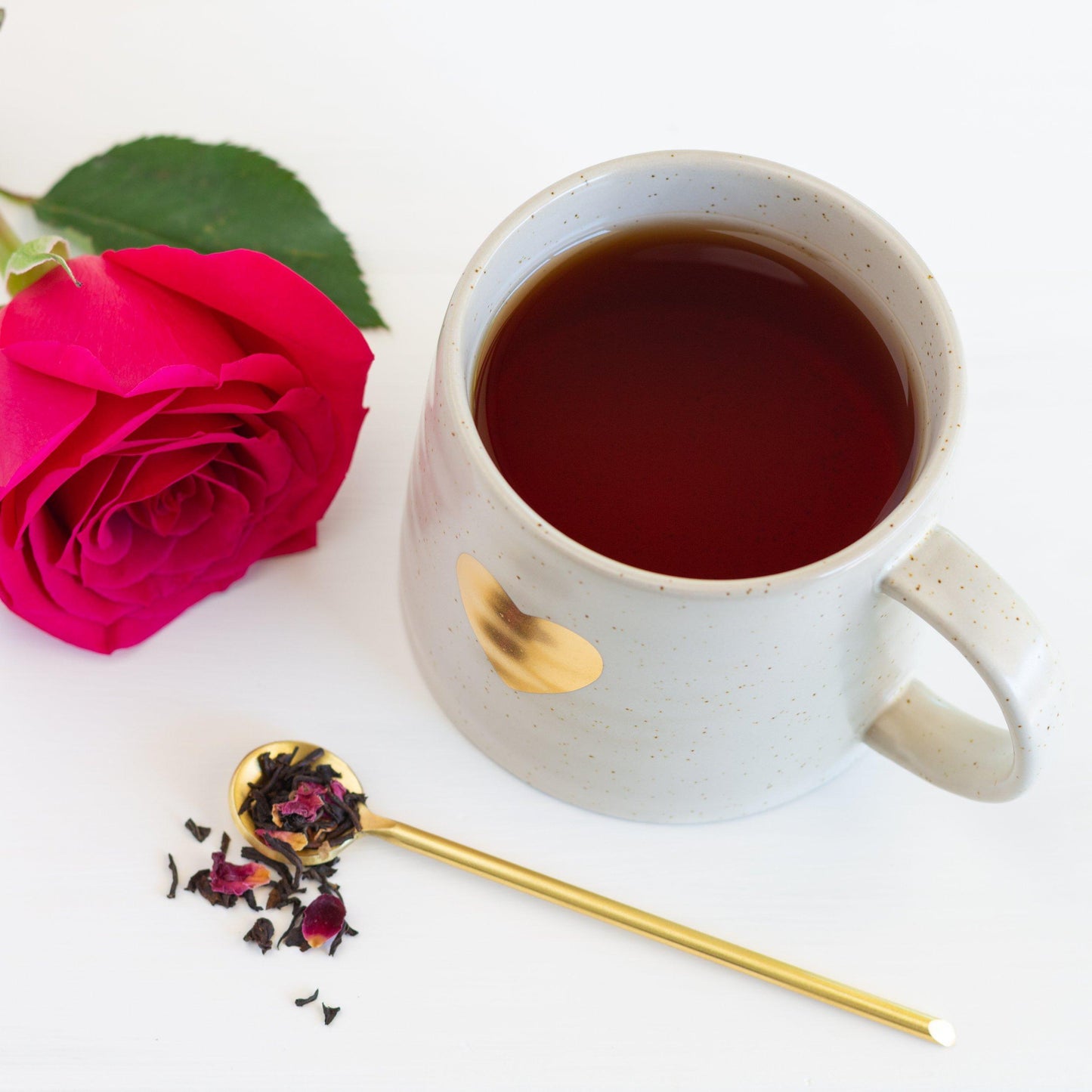 Midnight Rose Organic Black Tea shown as brewed tea in a white mug with a gold heart on it. Nearby is a dark pink rose and a thin gold spoon of tea leaves