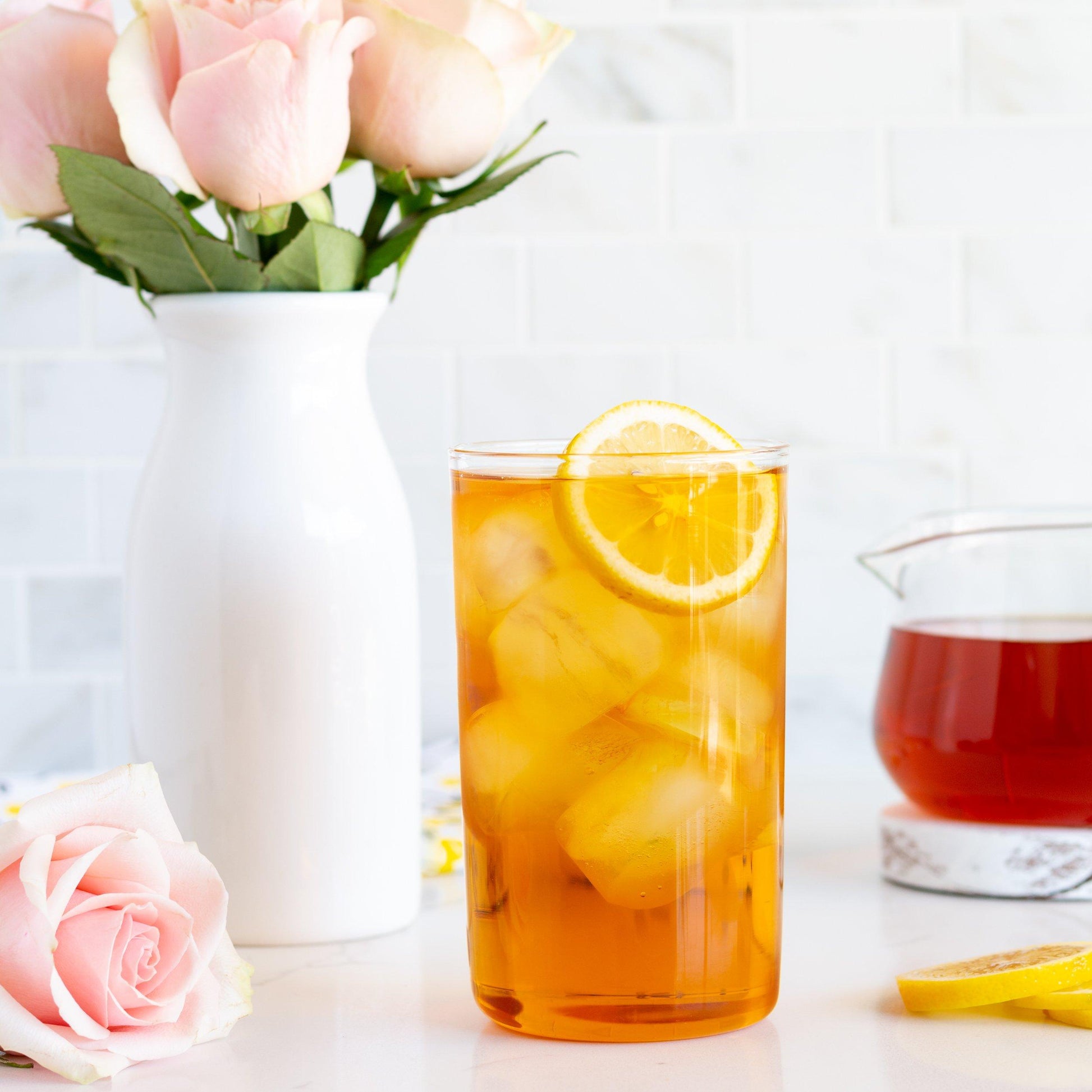 Midnight Rose Organic Black Tea shown as iced tea with a slice of lemon in a glass. Nearby is a white vase of pale pink roses, with a glass teapot of brewed tea in the background