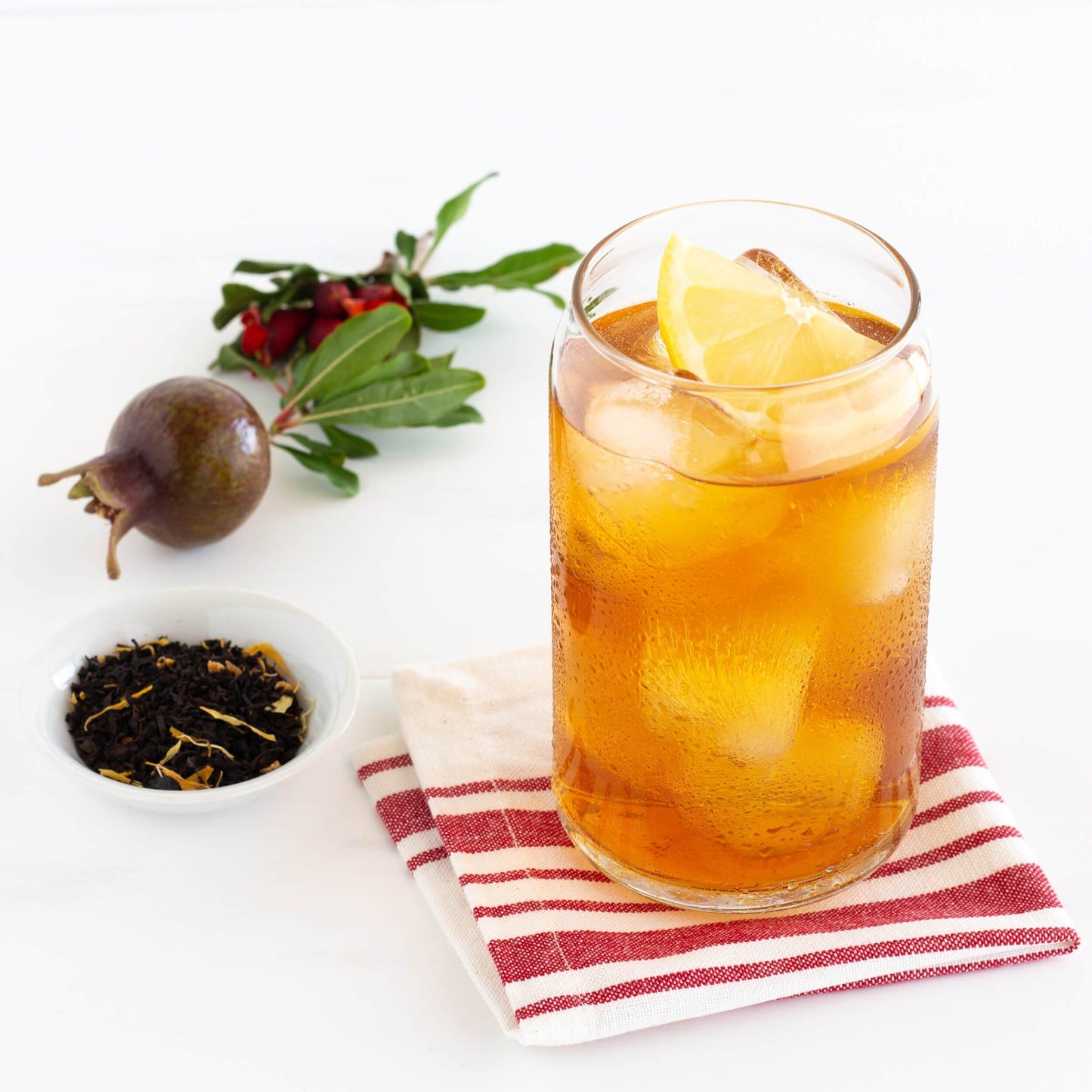 Pomegranate Lemon Organic Black Tea shown as iced tea in a glass displayed on a red and white striped napkin, with a small pomegranate and a tiny dish of loose tea leaves nearby