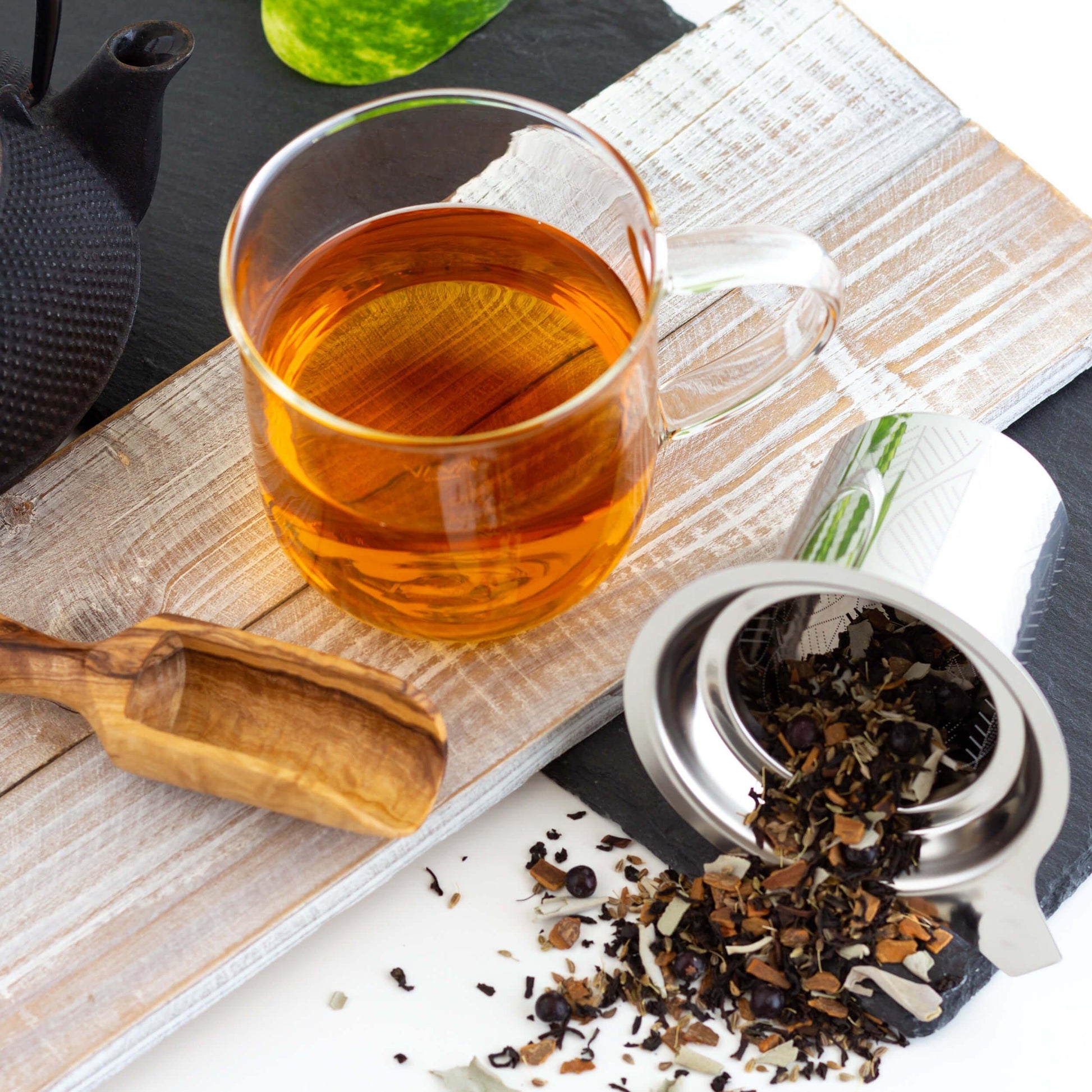 Santa Fe Sage Black Tea shown as brewed tea in a glass mug on a whitewashed wooden plank. A metal infuser is on its side with loose leaf tea spilling out, and a wooden scoop is nearby