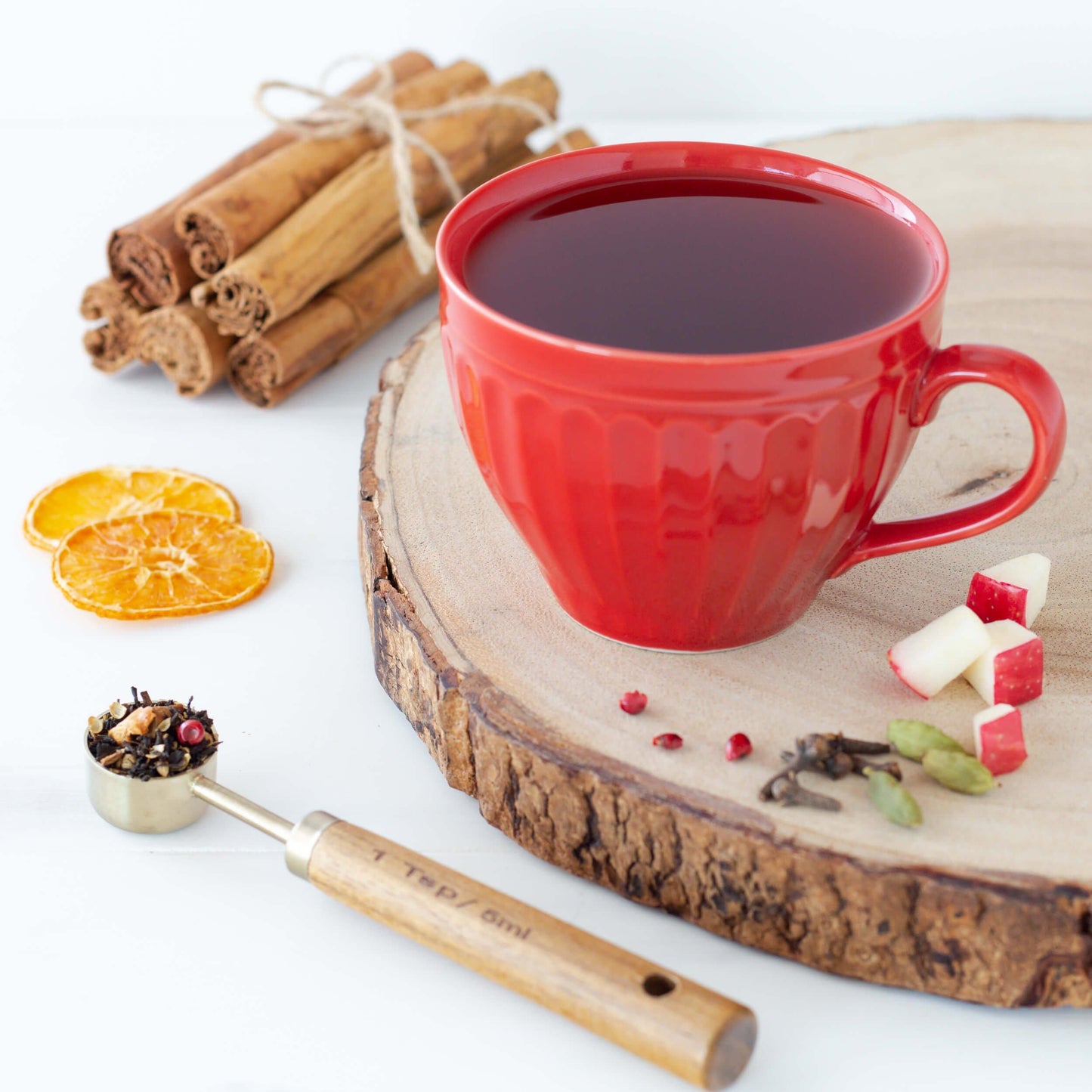 Solstice Spice Black Tea shown as brewed tea in a red cup displayed on a wooden disk, with orange slices, cinnamon sticks and other ingredients nearby