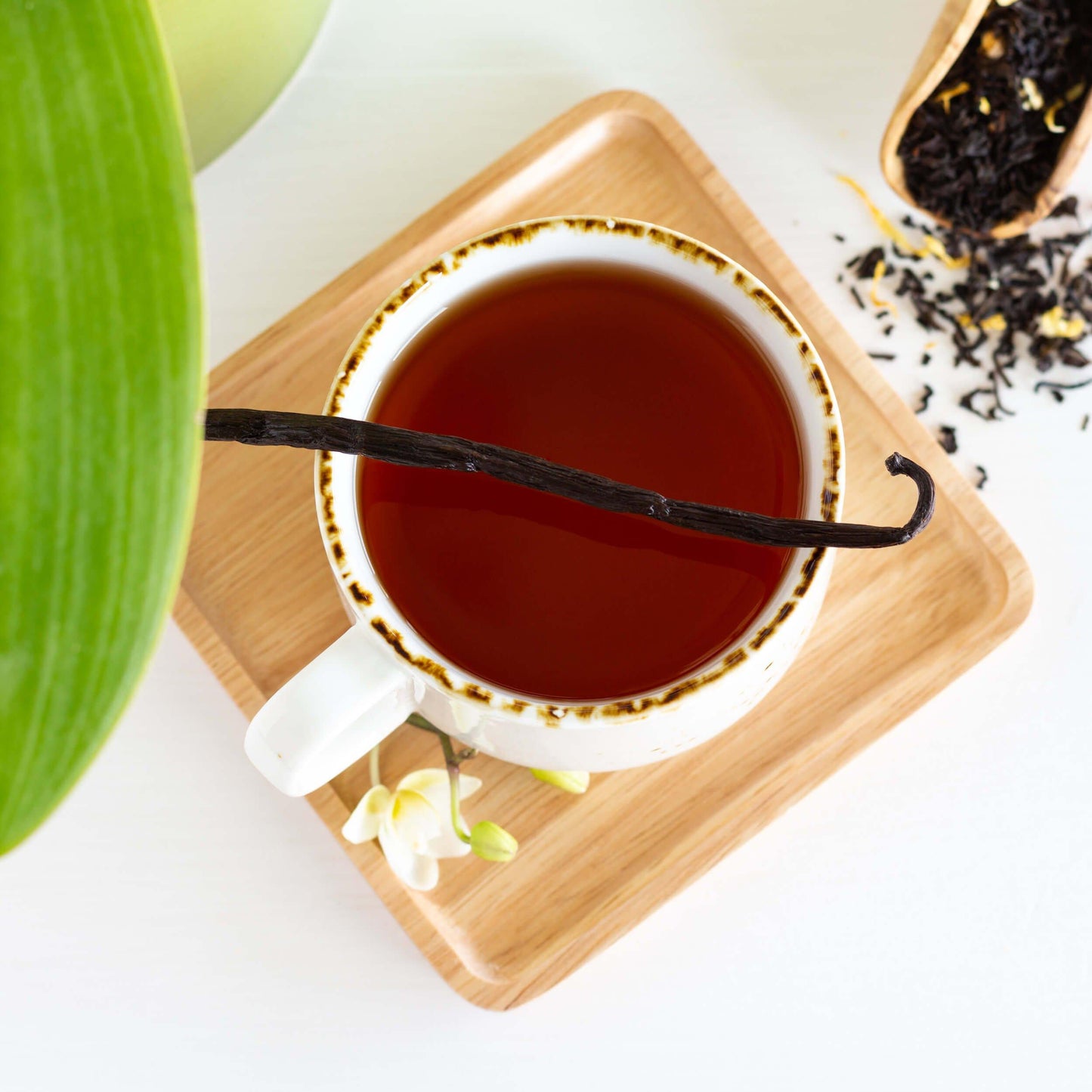 Vanilla Velvet Black Tea shown from above as brewed tea in a white mug with brown rim, displayed on a square bamboo coaster, with a vanilla bean lying across the top of the mug. 