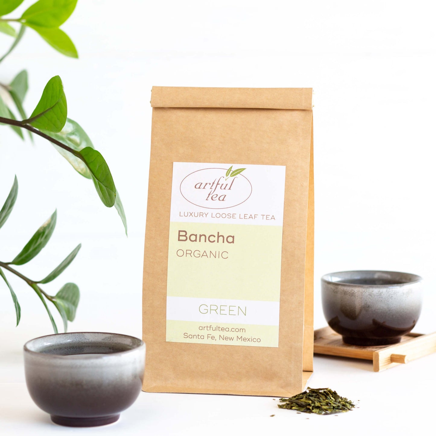 Banch Organic Green Tea shown packaged in a kraft bag, with two small black and gray teacups nearby. A green plant extends into the left side of the photo.