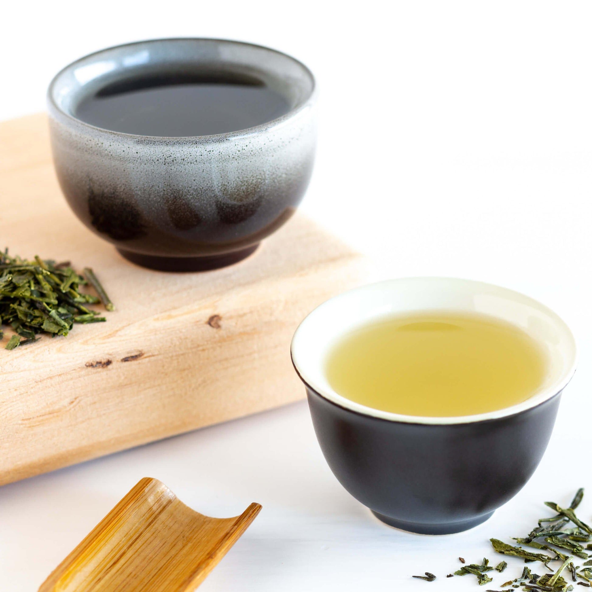 Bancha Organic Green Tea shown as brewed tea in two different small black teacups. One teacup has a gray glaze and is sitting on a wooden plank. Loose green tea leaves are nearby. A wooden scoop is in the foreground.