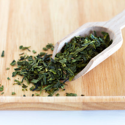 Bancha Organic Green Tea shown as loose tea leaves in close up spilling out of a small wooden scoop on a wooden tray.