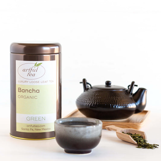 Bancha Organic Green Tea is shown packaged in a brown tin, with a small black and gray teacup in the foreground, a wooden scoop of loose tea leaves nearby, and a black teapot on a wooden tray in the background.