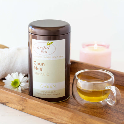 Chun Mee Organic Green Tea shown packaged in a brown tin, displayed on a wooden tray next to a tiny clear teacup of tea, a white daisy and a white cloth. A pink candle is lit in the background.