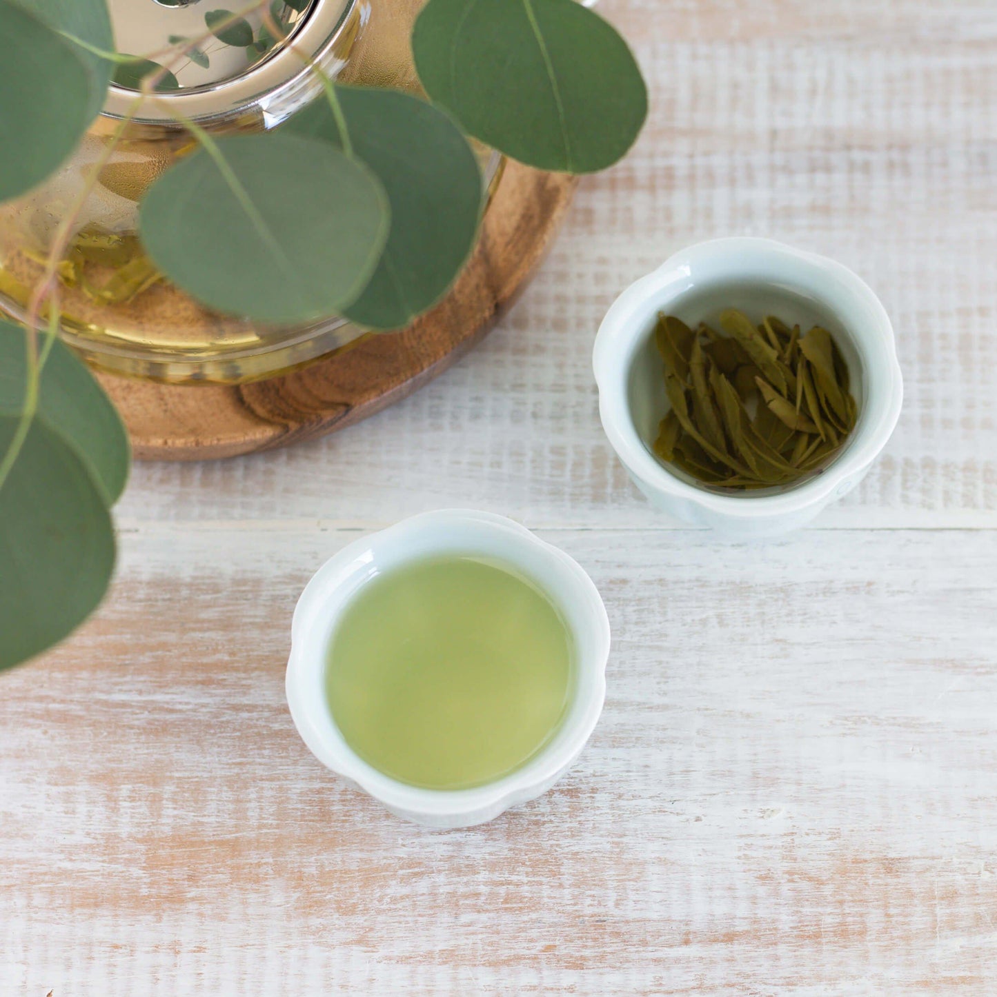 Dragon Well Superior Organic Green Tea shown as brewed tea in two small celadon teacups. One cup has many unfurled tea leaves in it. A glass teapot and greenery are nearby