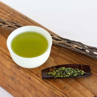 Genmaicha Matcha Organic Green Tea shown as brewed tea in a pale green teacup displayed on a wooden plank with a cherry wood scoop filled with loose leaf tea nearby