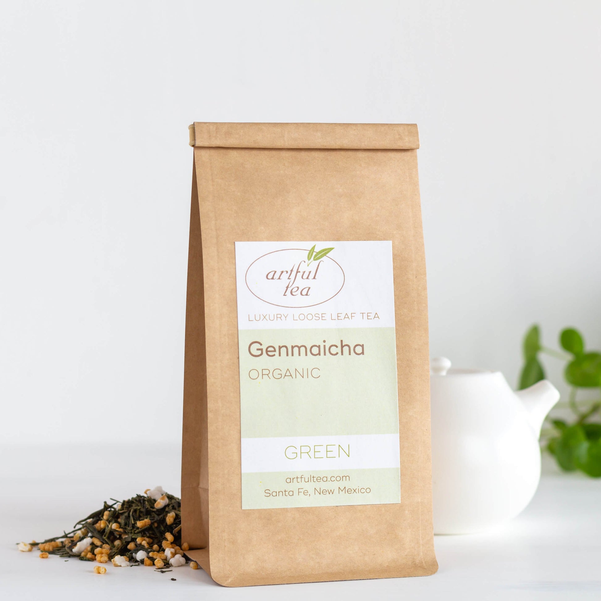 Genmaicha Organic Green Tea shown packaged in a kraft bag with loose tea leaves and a white teapot displayed behind the bag.