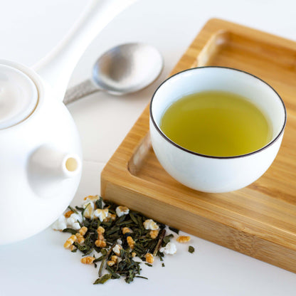 Genmaicha Organic Green Tea shown as brewed tea in a small white cup on a bamboo tray. A white teapot, a stainless steel scoop, and Genmaicha tea leaves surround the tray.