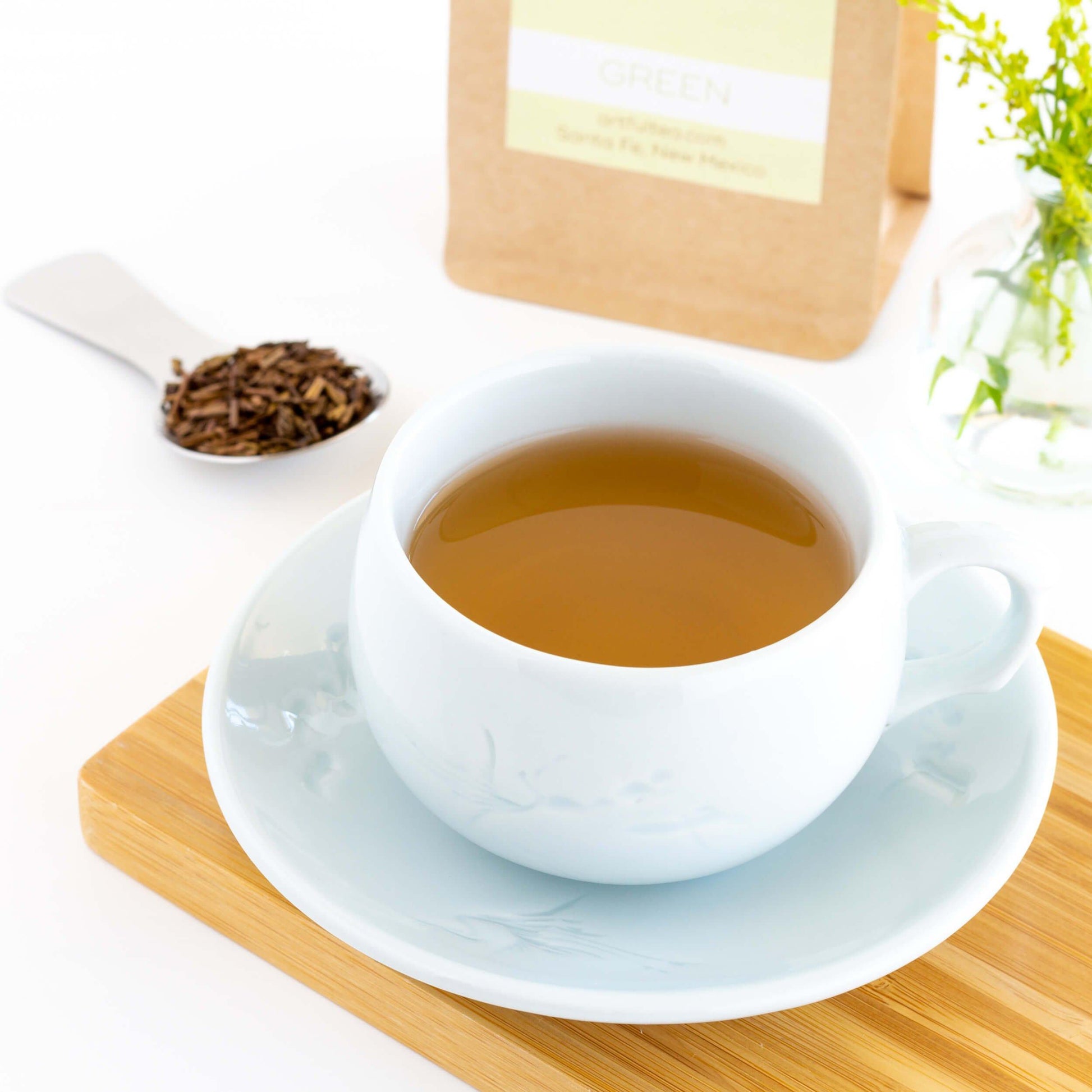 Hojicha Organic Green Tea shown as brewed tea in a pale blue teacup and saucer on a bamboo tray, with a silver scoop of tea leaves and a kraft bag of packaged tea in the background
