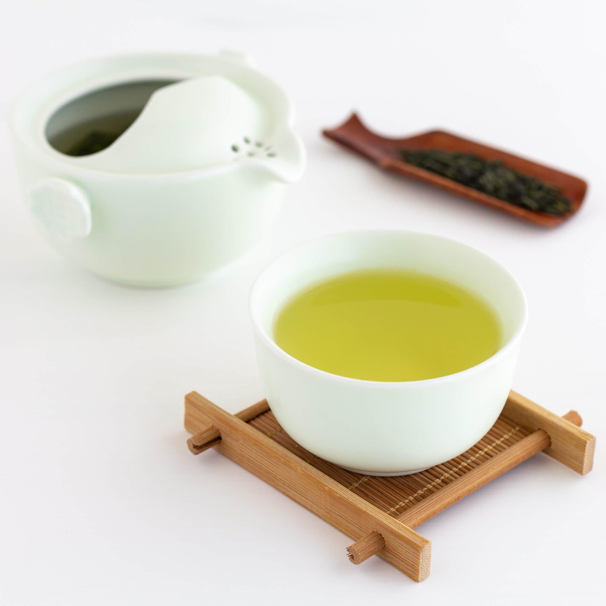 Kabusecha Organic Green Tea shown as brewed tea in a celadon teacup on  a bamboo coaster. Tiny celadon teapot and wooden scoop of loose leaf tea in the background