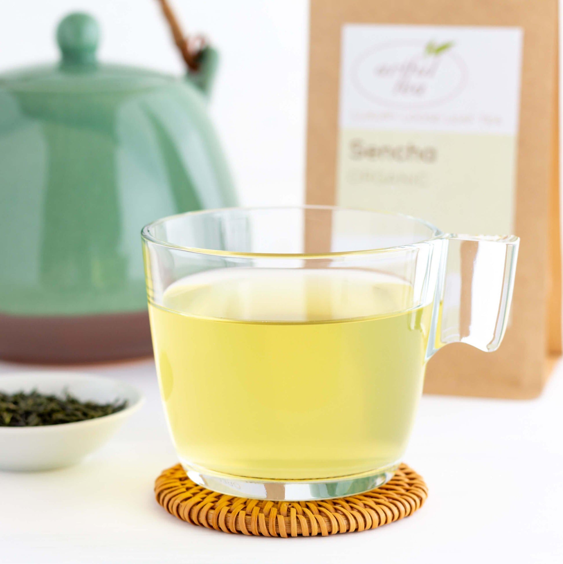 Sencha Organic Green Tea shown as brewed tea in a glass mug with teal green teapot and kraft bag of packaged tea in the background