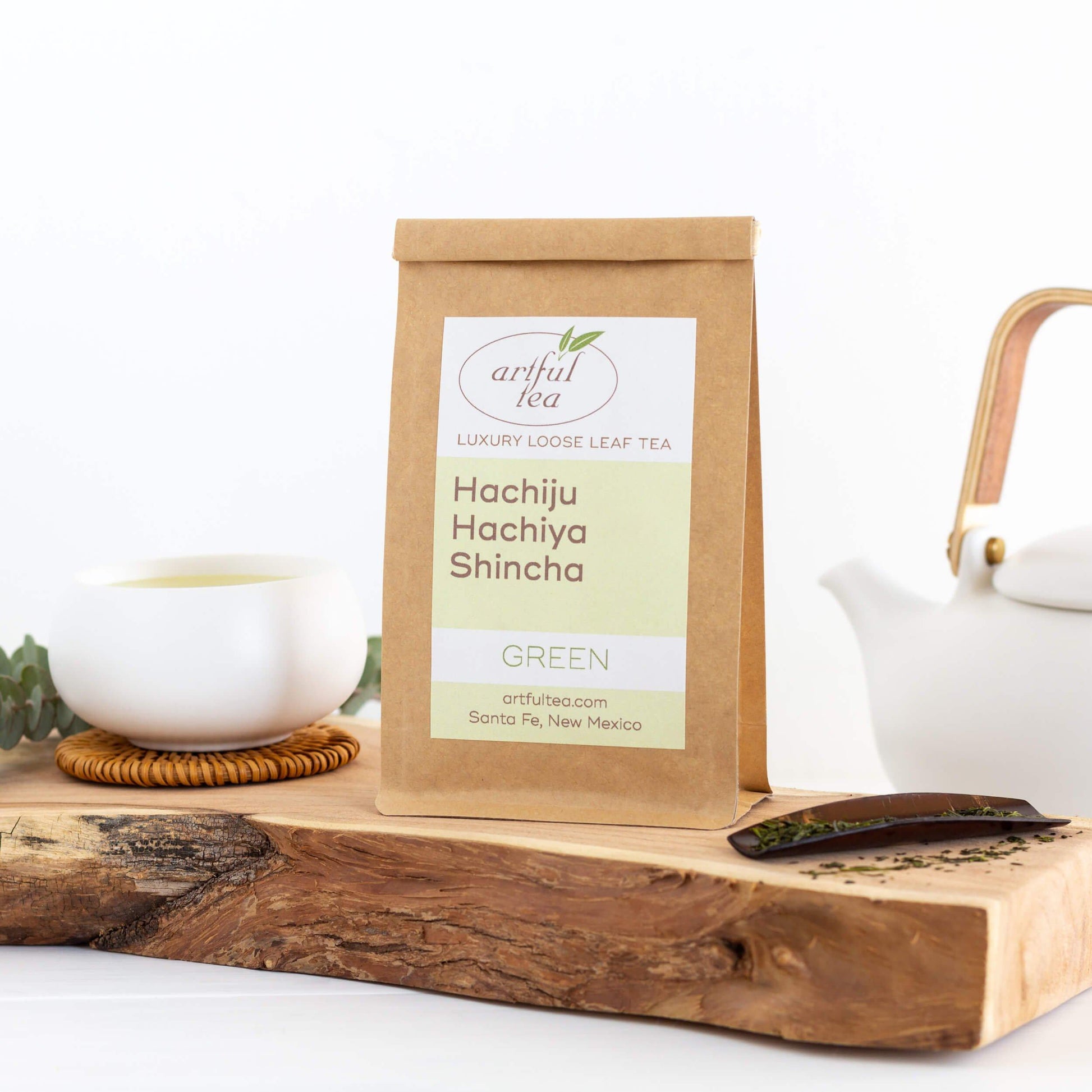 Shincha Green Tea shown packaged in a kraft bag, displayed on a wood plank next to a white cup. A white teapot is in the background.