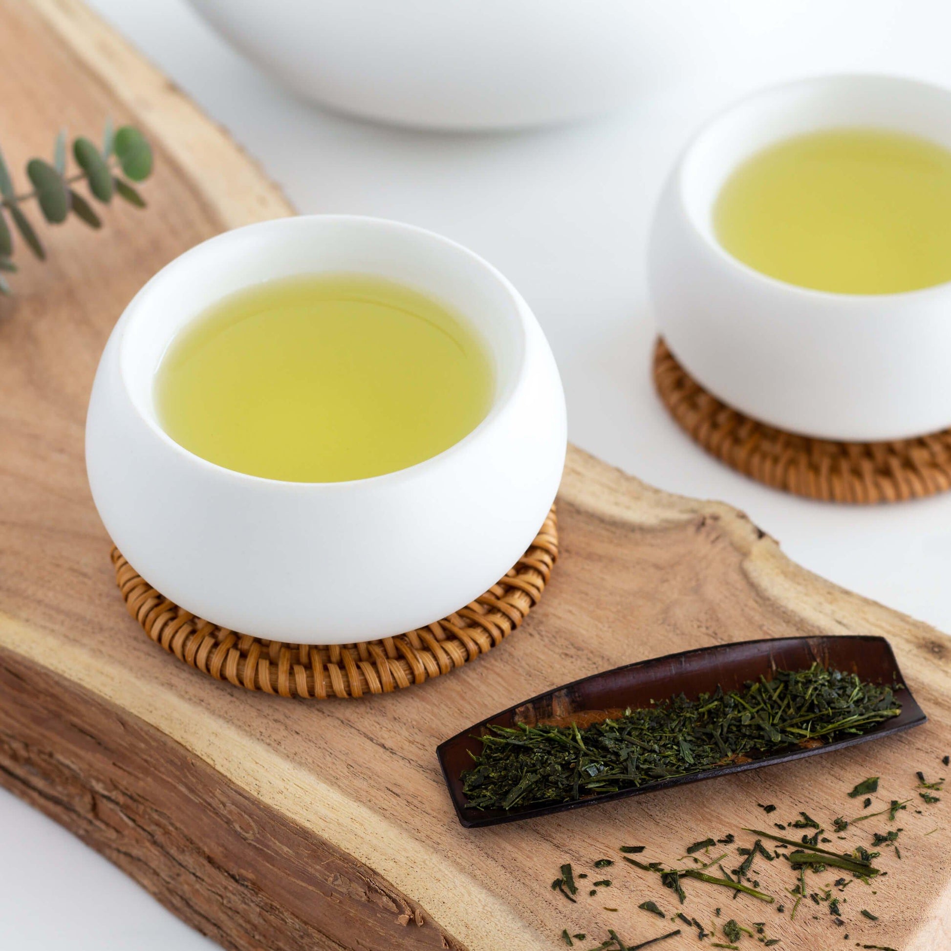 Shincha Green Tea shown as brewed tea in two small white cups, displayed on a wood plank with a Japanese cherry wood scoop with tea leaves in the foreground.