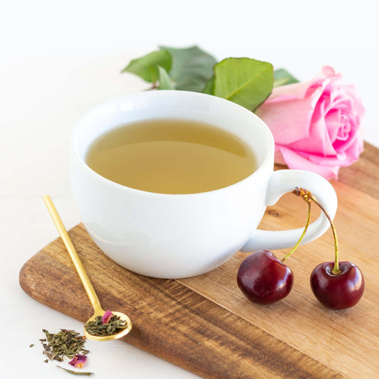 Cherry Rose Organic Green Tea shown as brewed tea in a white teacup displayed on a wood tray with a thin gold spoon, cherries and a pink rose