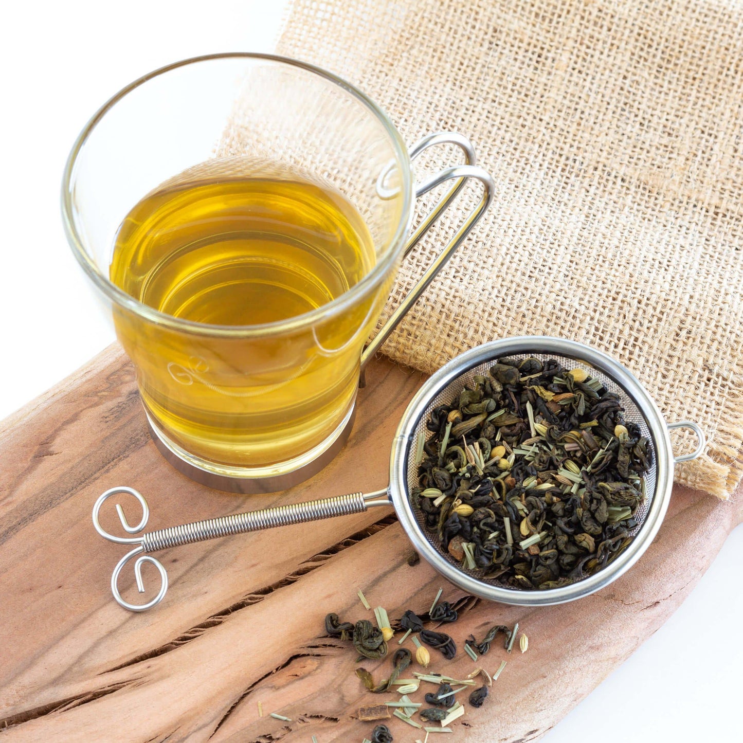 Green Tea Chai shown from above as brewed tea in a glass mug with a metal handle, displayed on a wood plank with burlap, and a strainer of loose tea leaves nearby