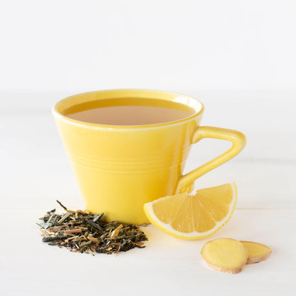 Linda’s Lemon Ginger Green Tea shown as brewed tea in a bright yellow mug, surrounded with a lemon wedge, ginger root slices, and loose tea leaves