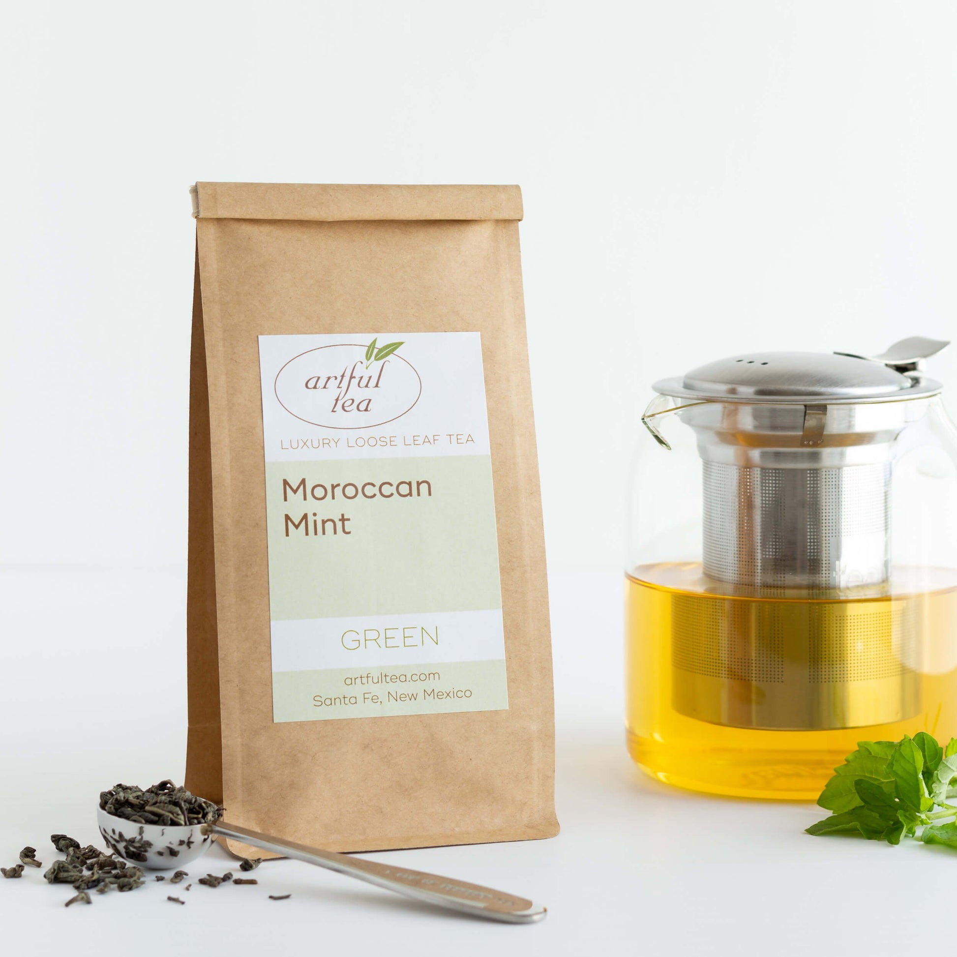 Moroccan Mint Green Tea shown packaged in a kraft bag with a scoop of loose tea leaves in the foreground. A glass teapot of brewing tea is nearby with some mint leaves next to it
