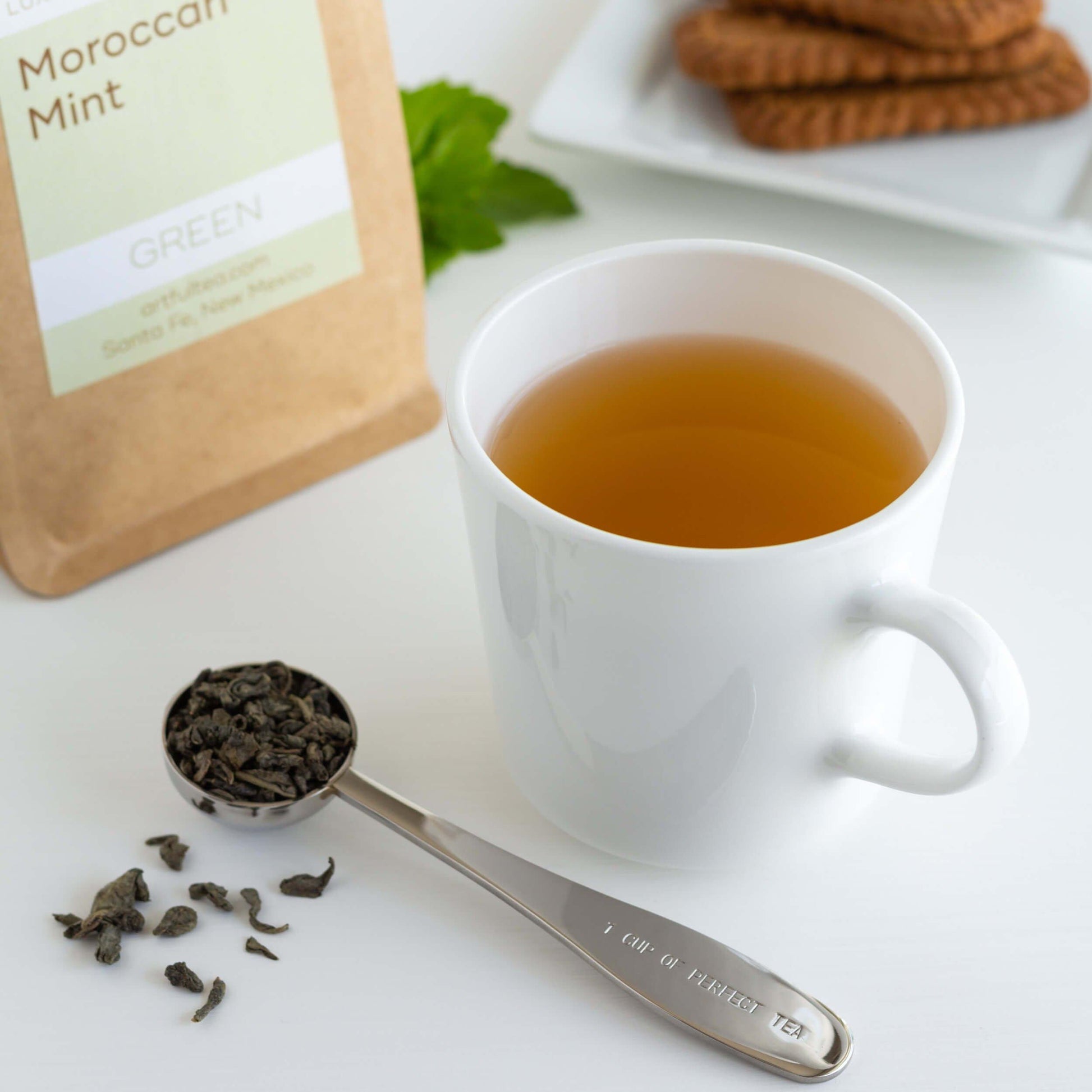 Moroccan Mint Green Tea shown as brewed tea in a white cup with a scoop of loose tea leaves nearby. A kraft bag of packaged tea and three cookies are on a white dish in the background