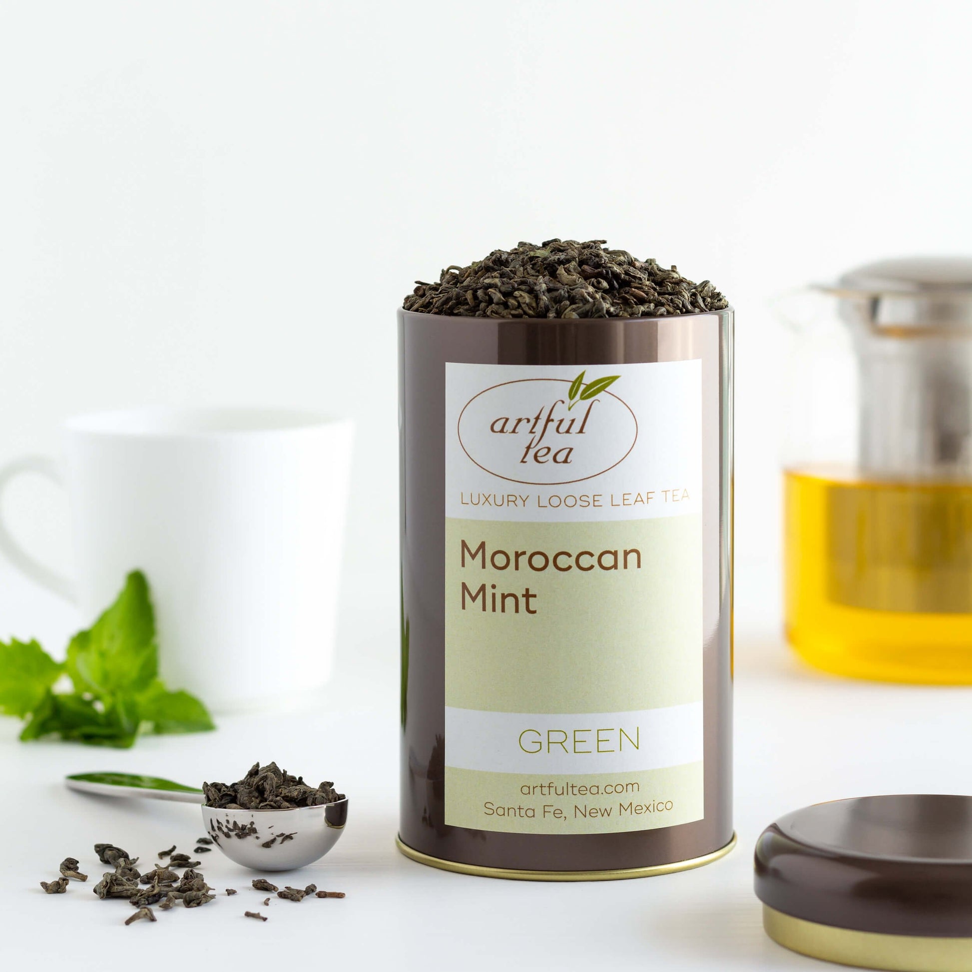 Moroccan Mint Green Tea shown packaged in a brown tin with the lid off, next to a tea scoop of loose tea leaves. A glass teapot and a white mug with mint leaves are in the background