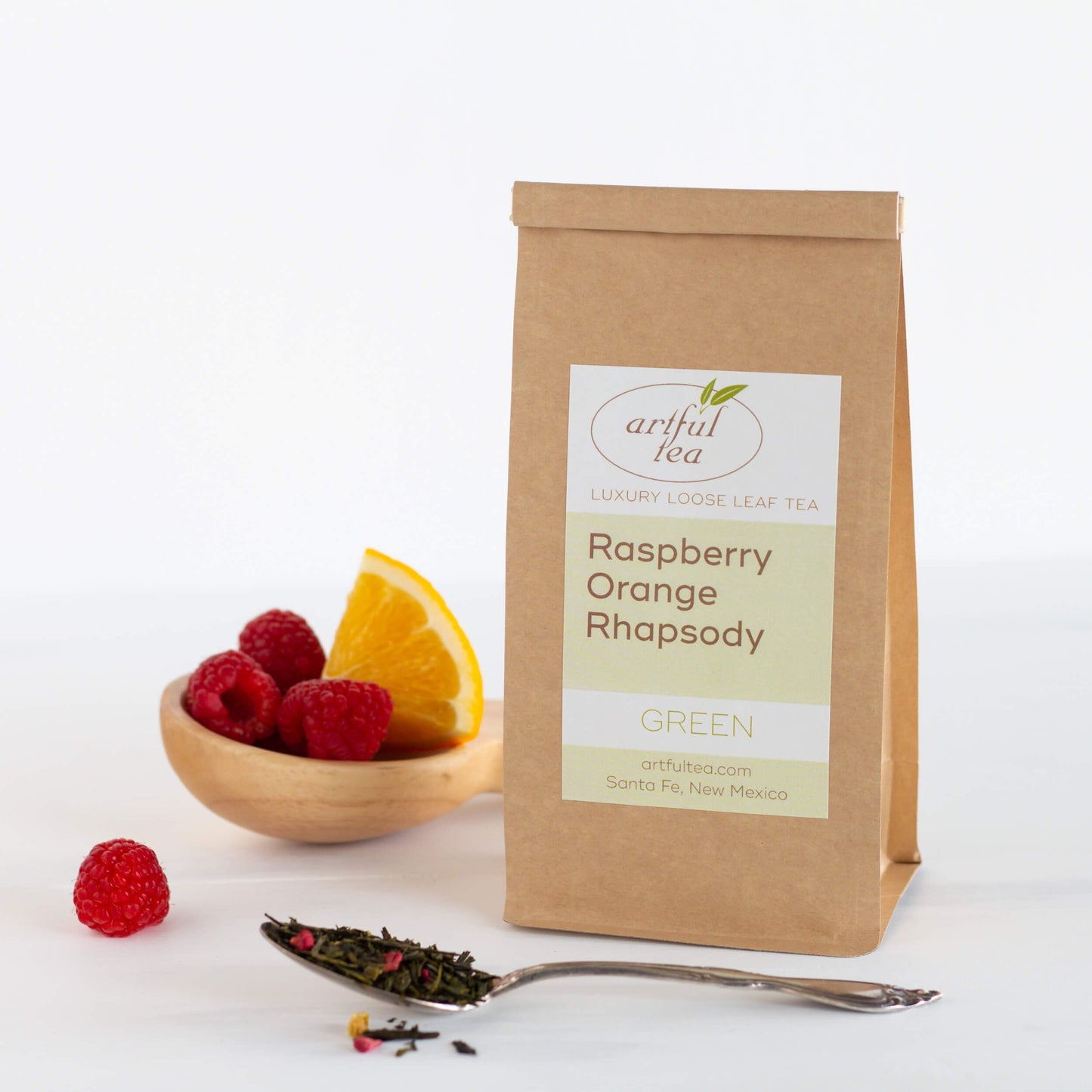 Raspberry Orange Rhapsody Green Tea shown packaged in a kraft bag, with a spoon full of tea leaves in the foreground and a wooden scoop with raspberries and an orange wedge displayed behind