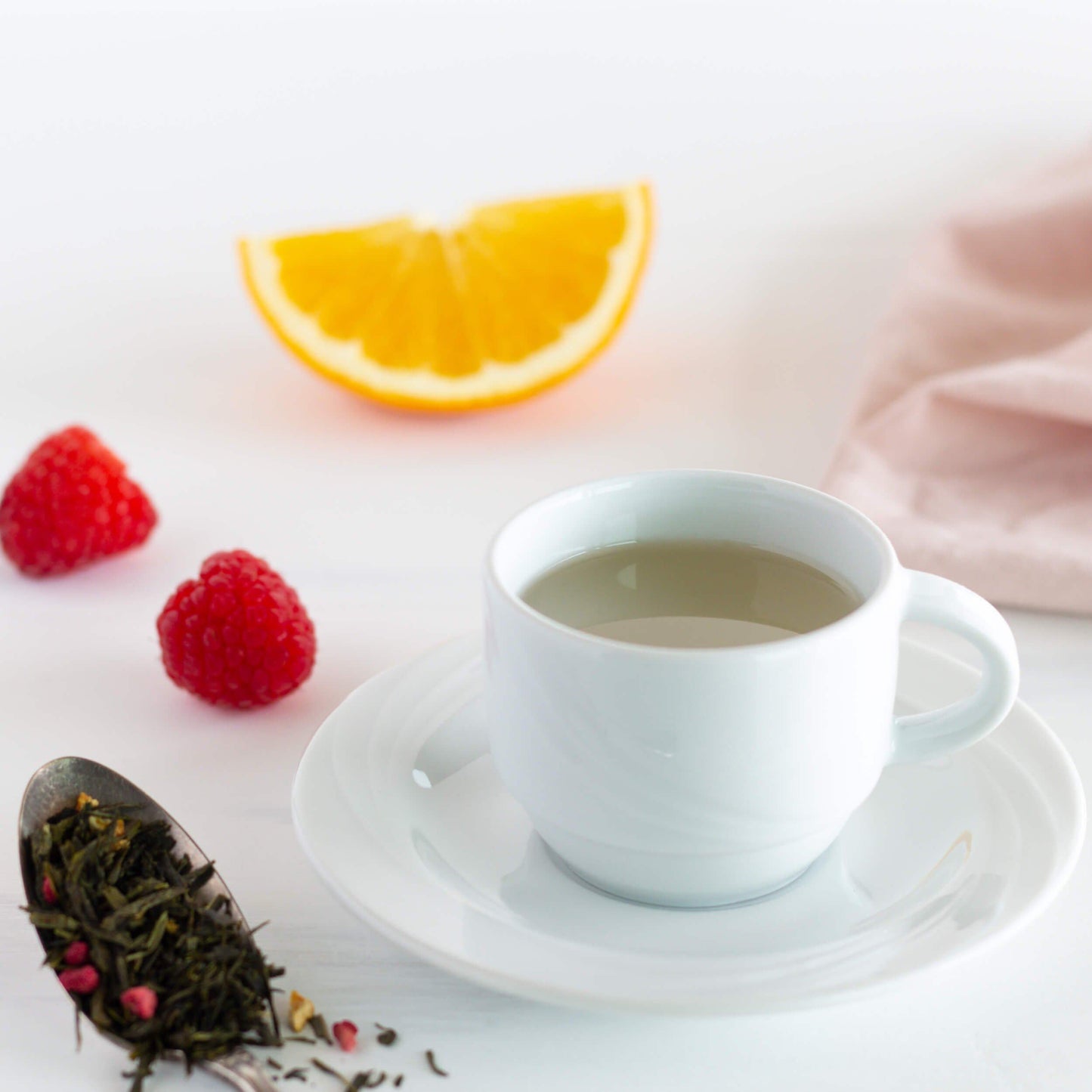 Raspberry Orange Rhapsody Green Tea shown as brewed tea in a white teacup and saucer with a wedge of orange and raspberries nearby