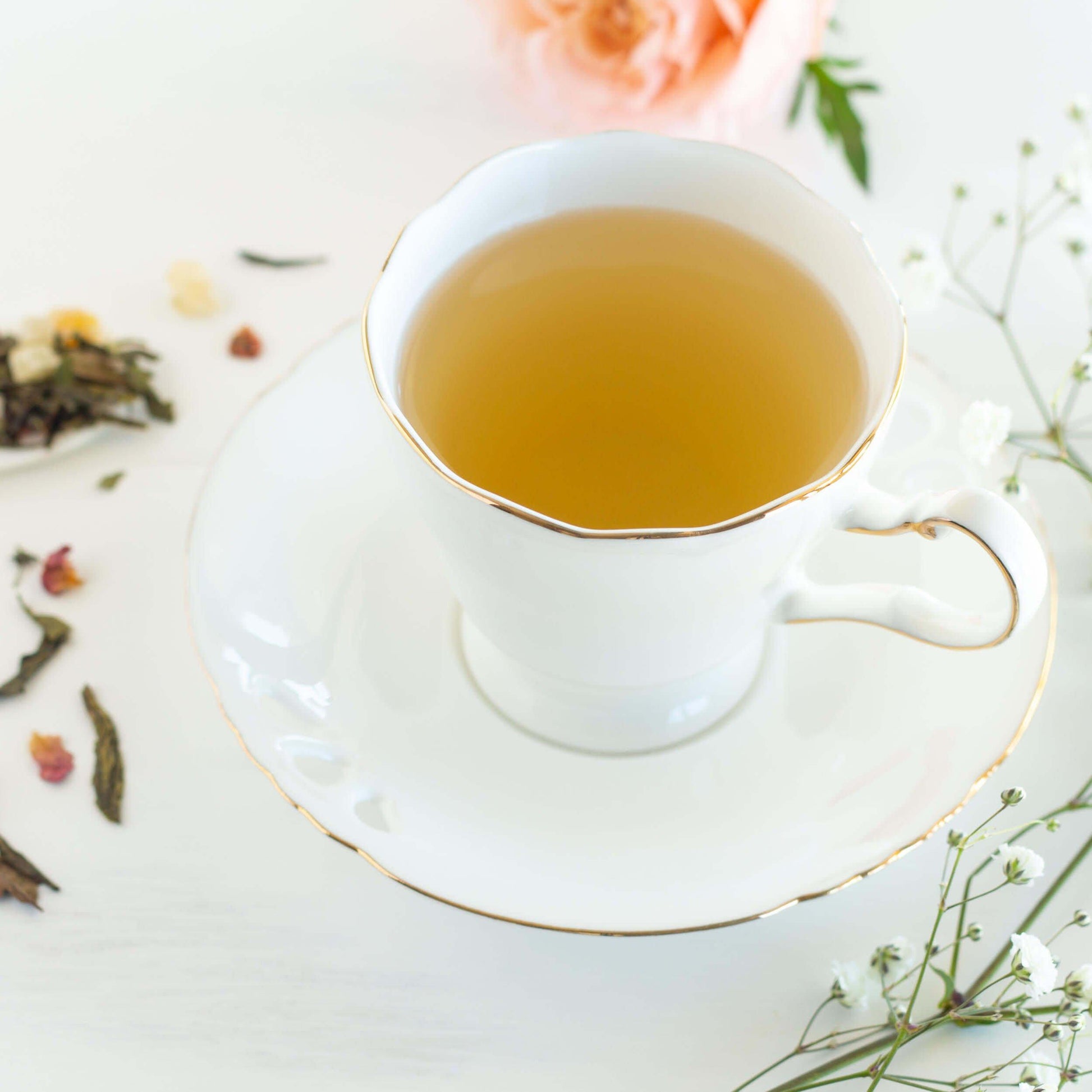 Spring Fancy Green and White Tea blend shown as brewed tea in a traditional white teacup and saucer with gold trim. Cup is surrounded by baby's breath, a pink rose, and loose tea leaves