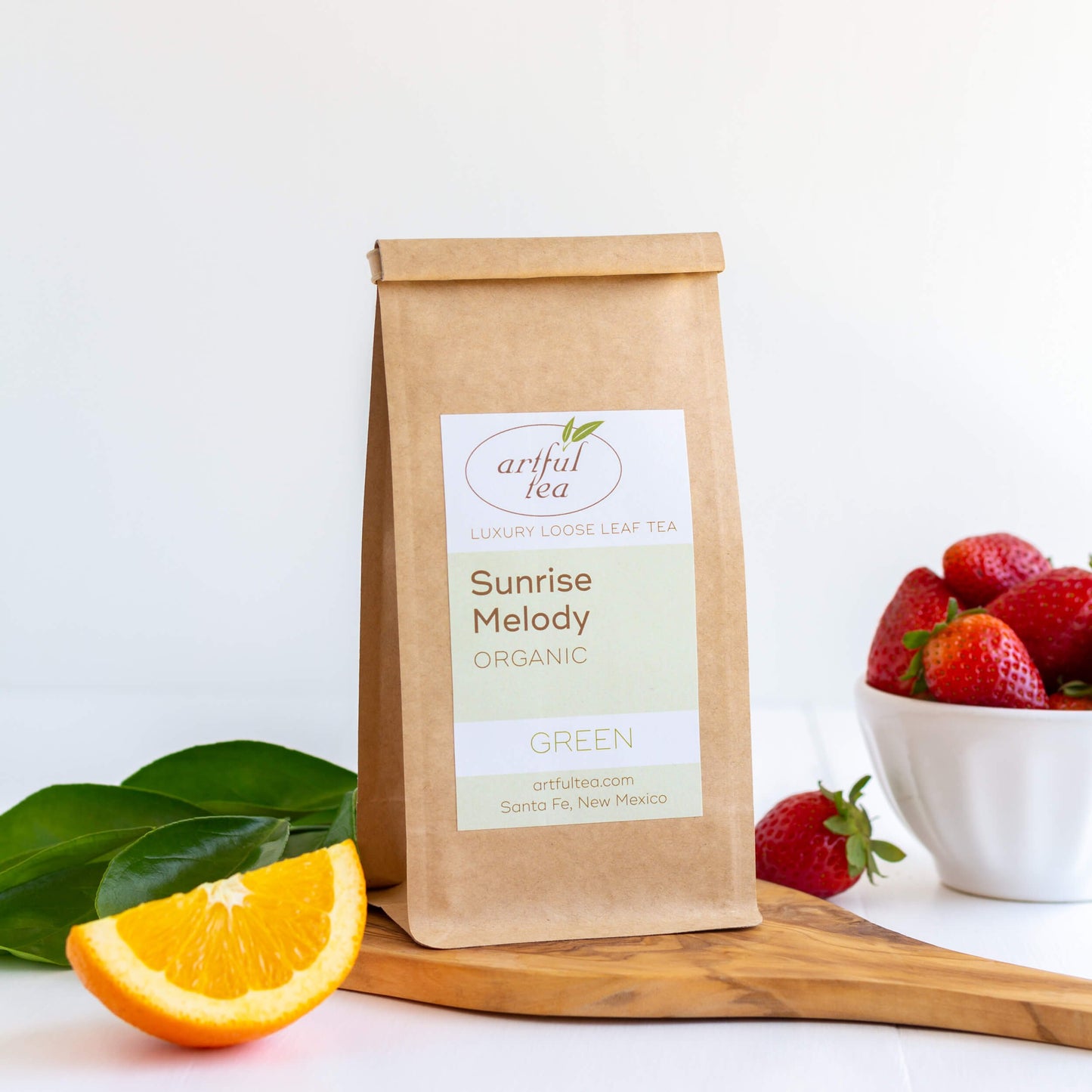 Sunrise Melody Organic Green Tea shown packaged in a kraft bag, displayed on a wooden board with a wedge of orange and a bowl of strawberries.