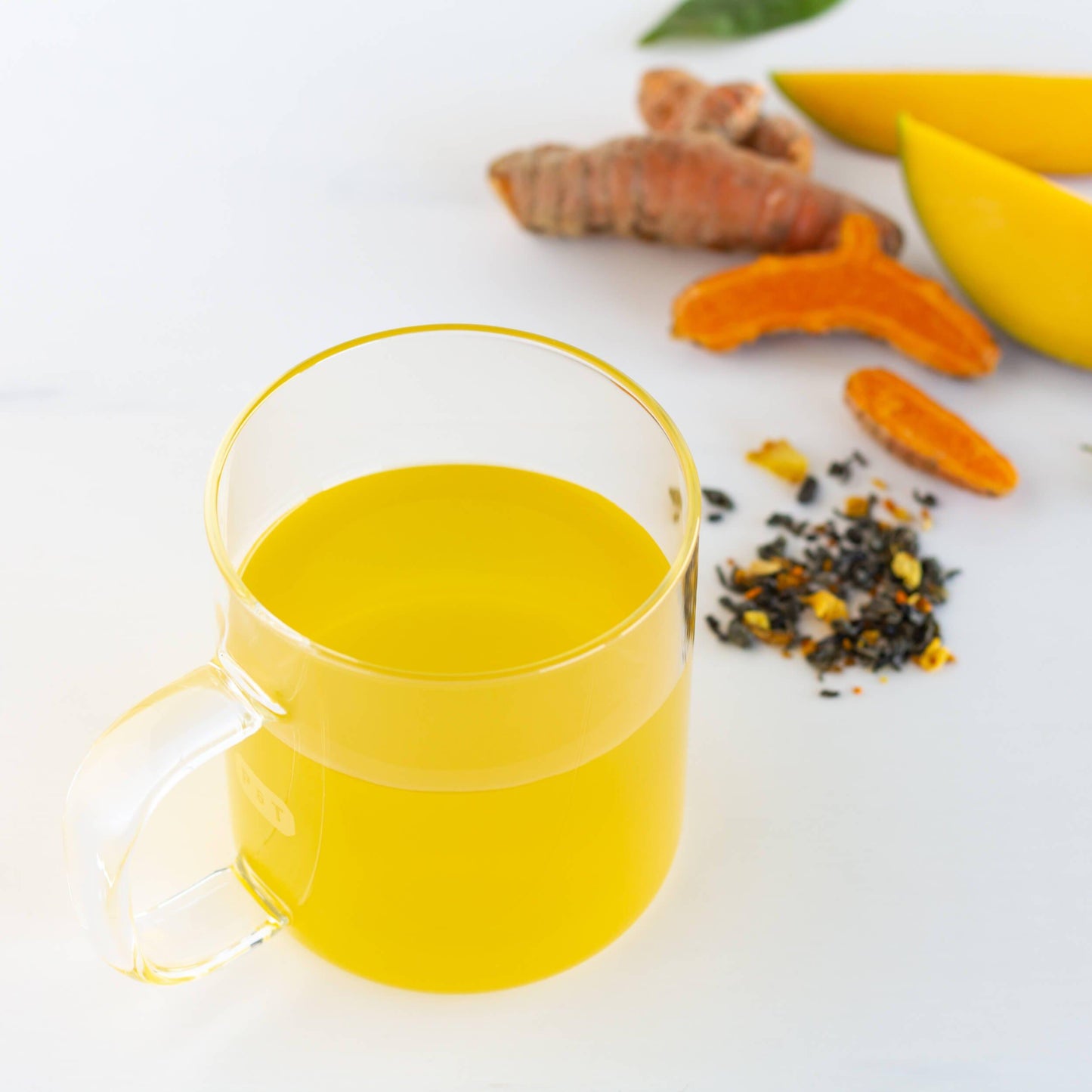 Turmeric Mango Organic Green Tea shown as brewed tea in a glass mug with turmeric root, sliced mango, and loose tea leaves in the background