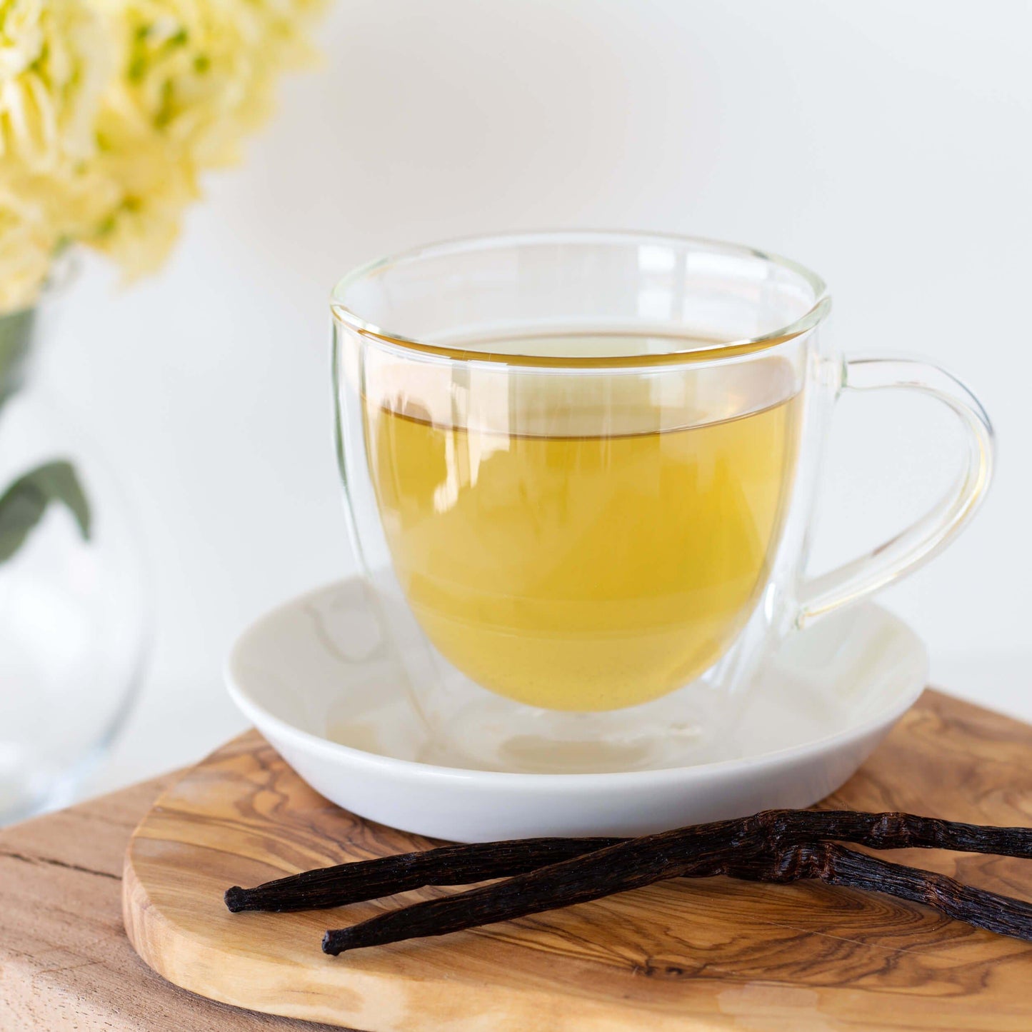 Vanilla Bean Organic Green Tea shown as brewed tea in a glass mug displayed in a white saucer on a wooden board, with two vanilla beans in the foreground