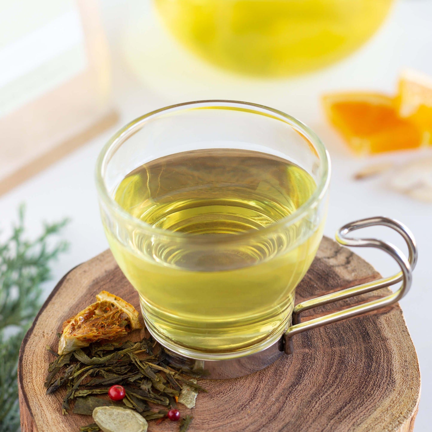 Winter Forest Green Tea shown as brewed tea in a glass mug with a metal handle, displayed on a wooden coaster with loose tea leaves. Orange slices and brewed tea are in the background