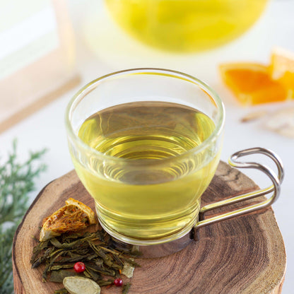 Winter Forest Green Tea shown as brewed tea in a glass mug with a metal handle, displayed on a wooden coaster with loose tea leaves. Orange slices and brewed tea are in the background