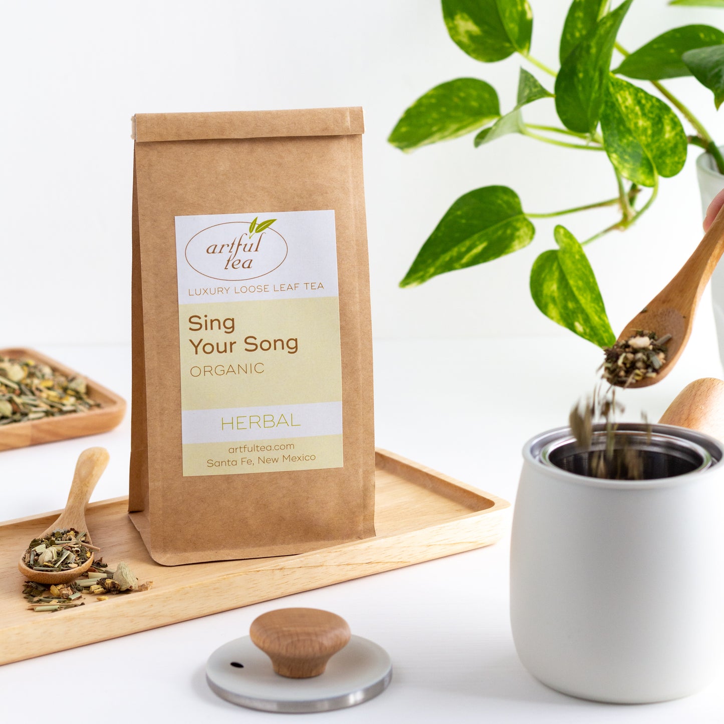 Organic Sing Your Song Herbal Tea shown packaged in a kraft bag, displayed on a wooden tray. Tea leaves are being spooned into a white teapot in the foreground. A green plant is in the background.