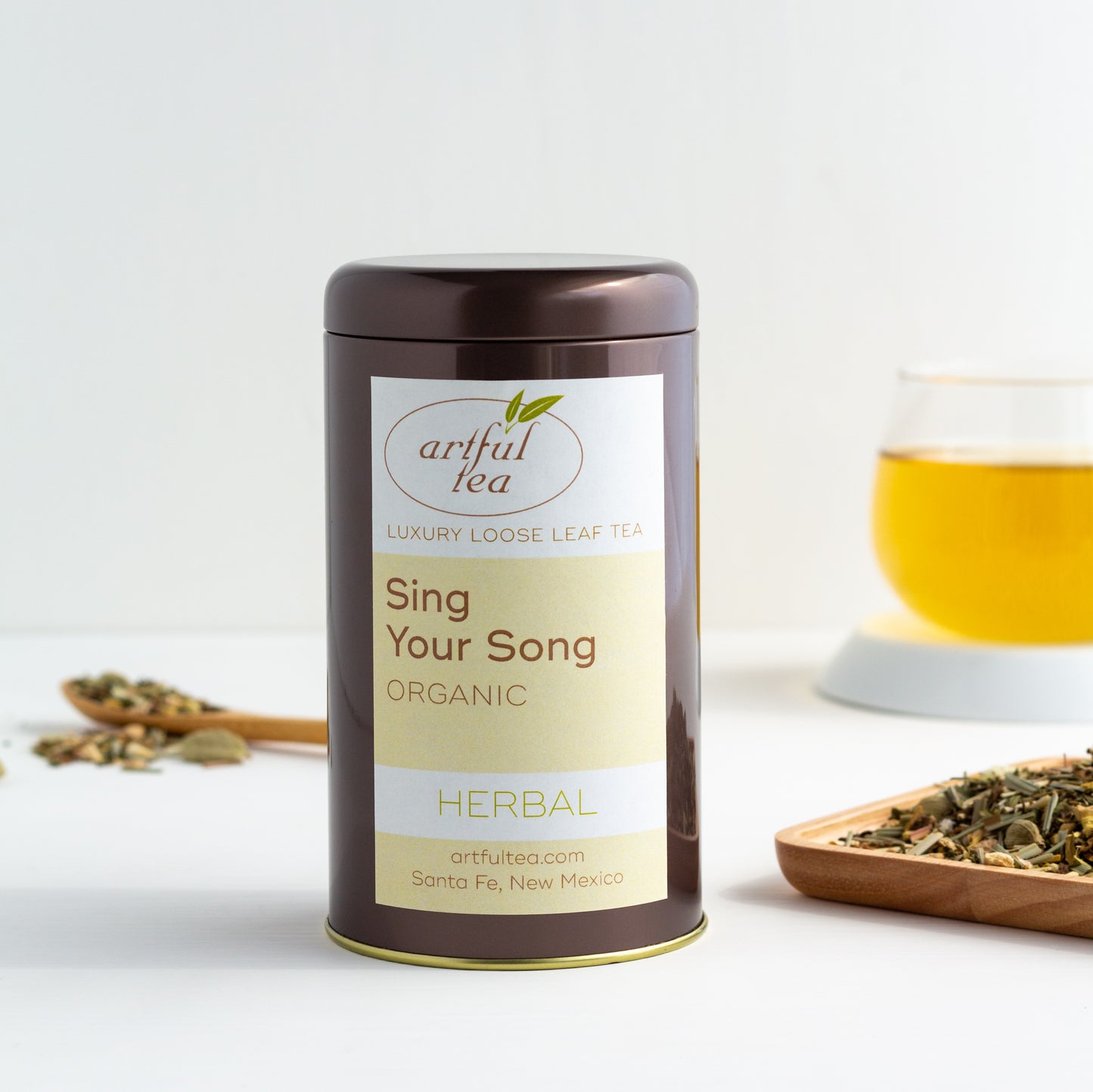 Organic Sing Your Song Herbal Tea shown packaged in a brown tin with a wooden dish of tea leaves nearby and a glass of brewed tea in the background.
