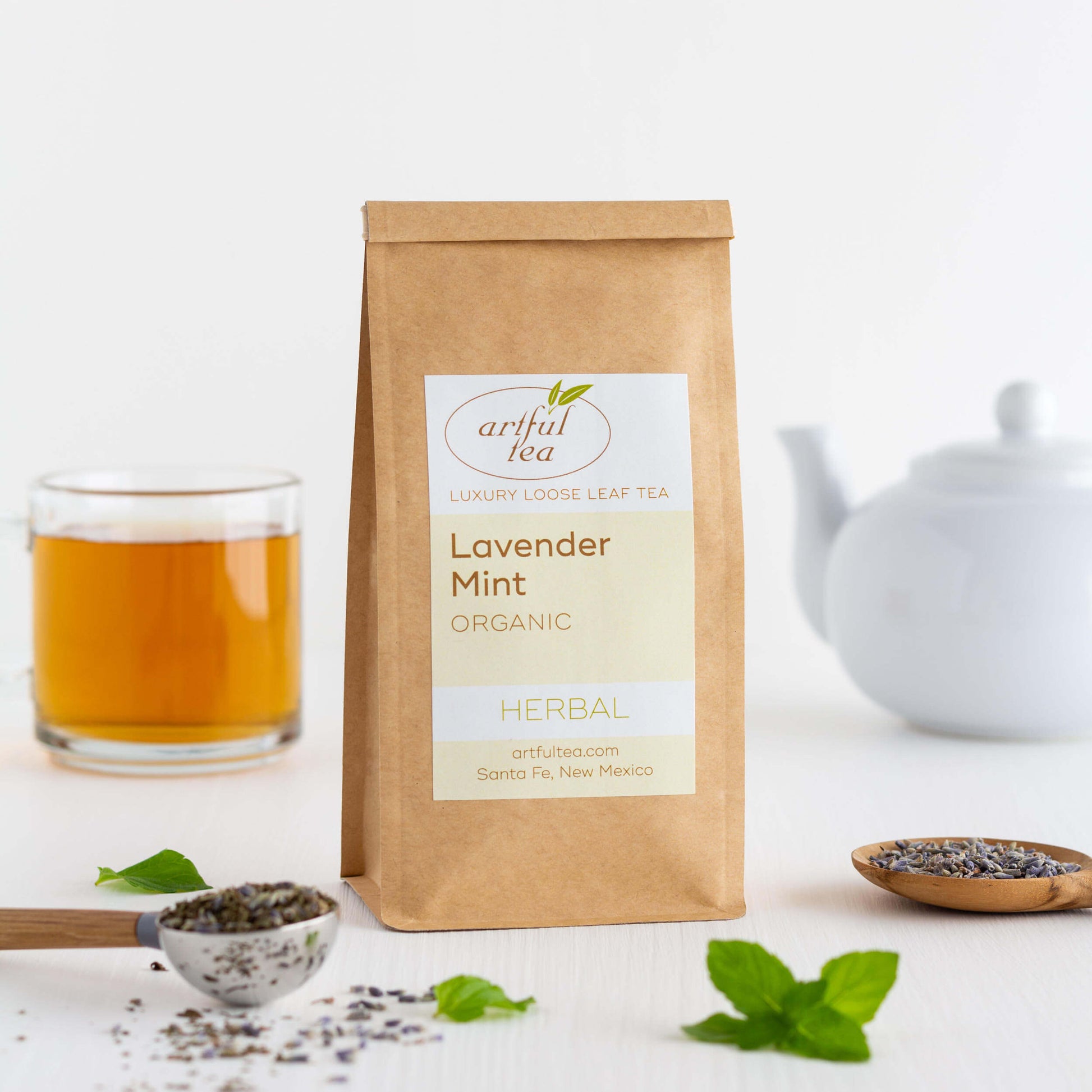 Lavender Mint Organic Herbal Tea shown packaged in a kraft bag with a scoop of tea leaves and some fresh mint leaves in the foreground. A glass mug of brewed tea and a white teapot are in the background