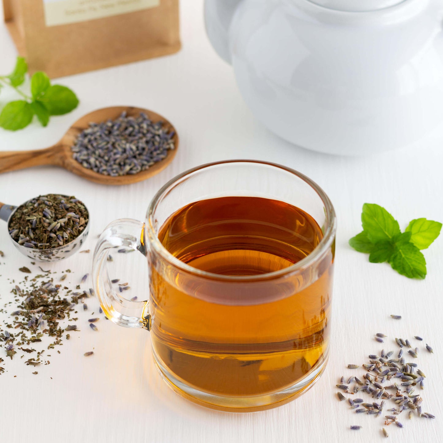 Lavender Mint Organic Herbal Tea shown as brewed tea in a glass mug surrounded by loose tea leaves and loose lavender buds. A white teapot is in the background