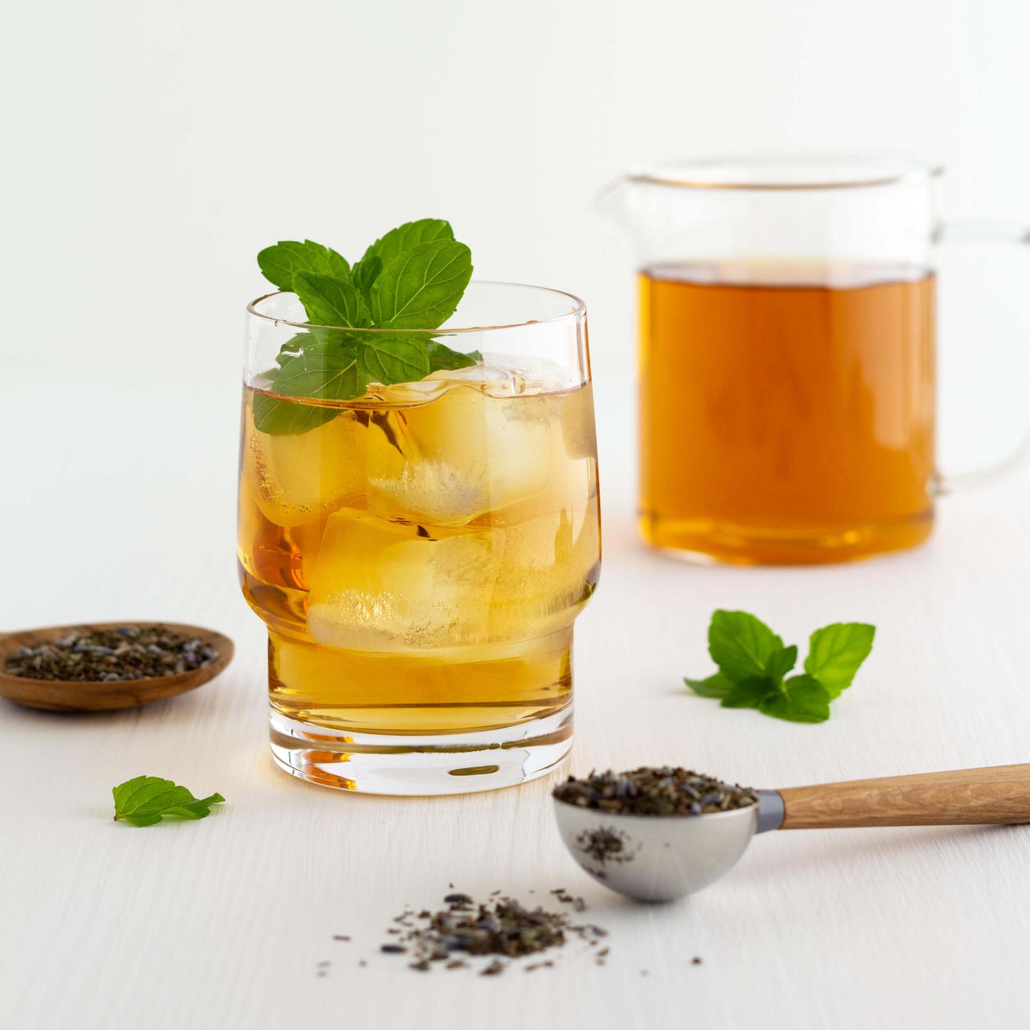 Lavender Mint Organic Herbal Tea shown as iced tea in a short glass with a sprig of mint leaves. A scoop of loose tea leaves is in the foreground. A glass pitcher of brewed tea is in the background