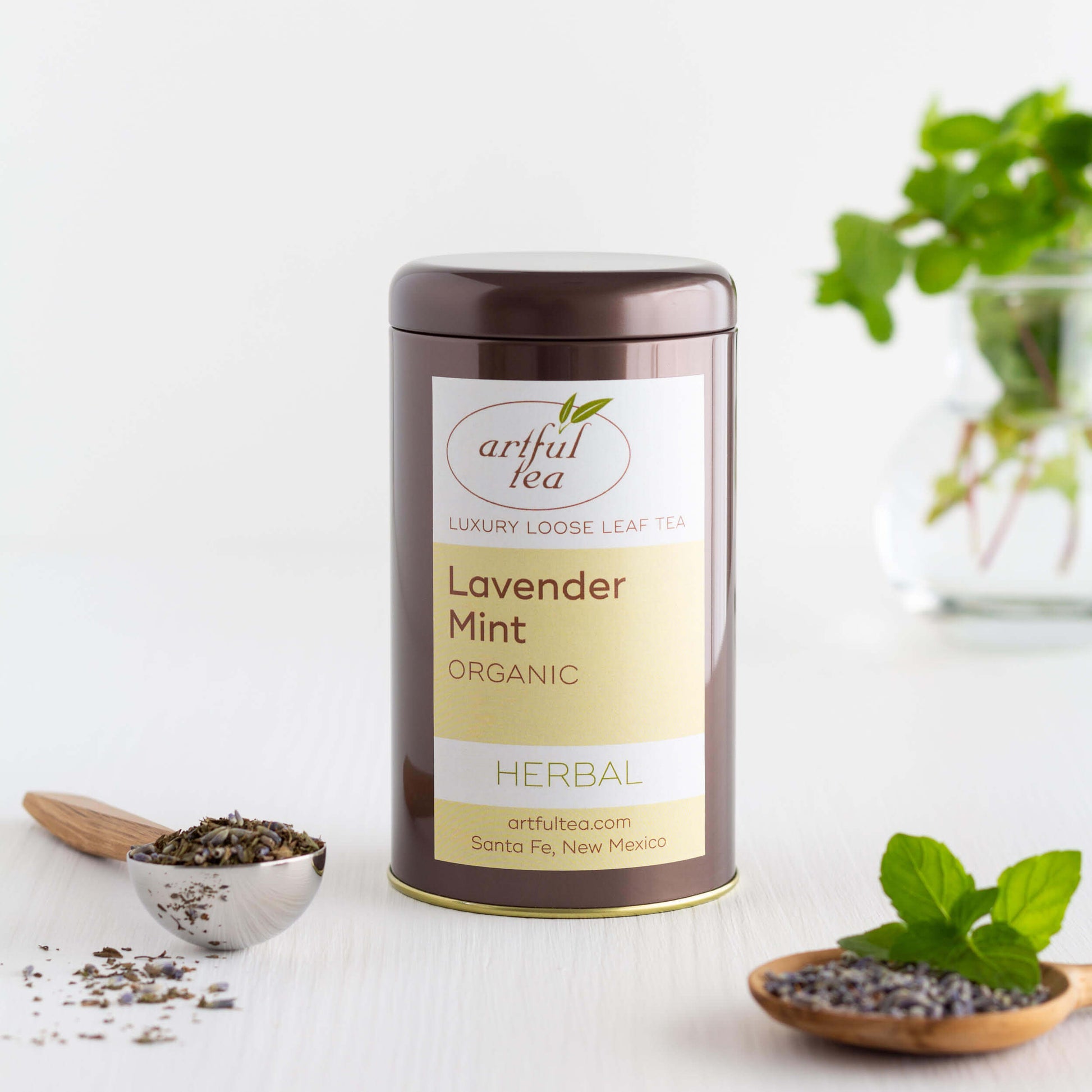 Lavender Mint Organic Herbal Tea shown packaged in a brown tin, with a scoop of loose tea leaves in the foreground and a glass vase with sprigs of mint in the background