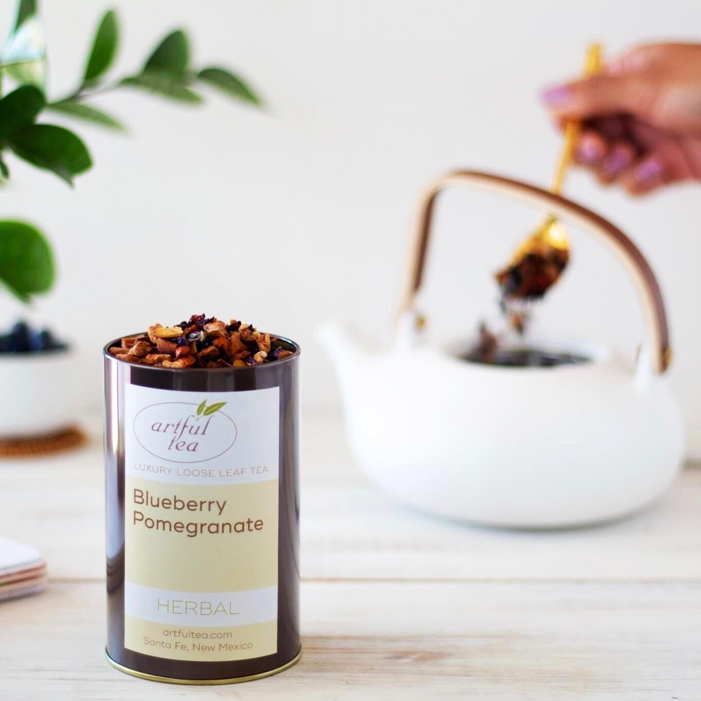 Blueberry Pomegranate Herbal Tea displayed in an open brown tin. In the background, a hand is spooning tea into a white teapot