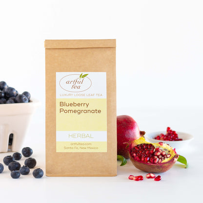 Blueberry Pomegranate Herbal Tea shown packaged in a kraft bag with fresh blueberries and an open pomegranate displayed on either side