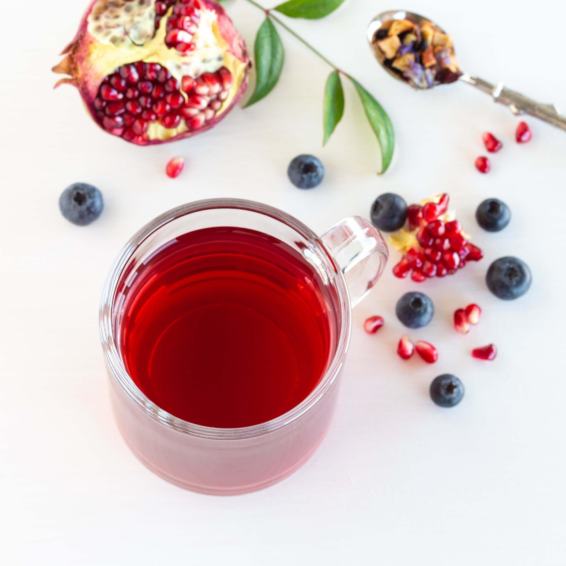 Blueberry Pomegranate Herbal Tea shown from above as brewed tea in a glass mug, surrounded by loose  blueberries, an open pomegranate, and a spoon of loose tea