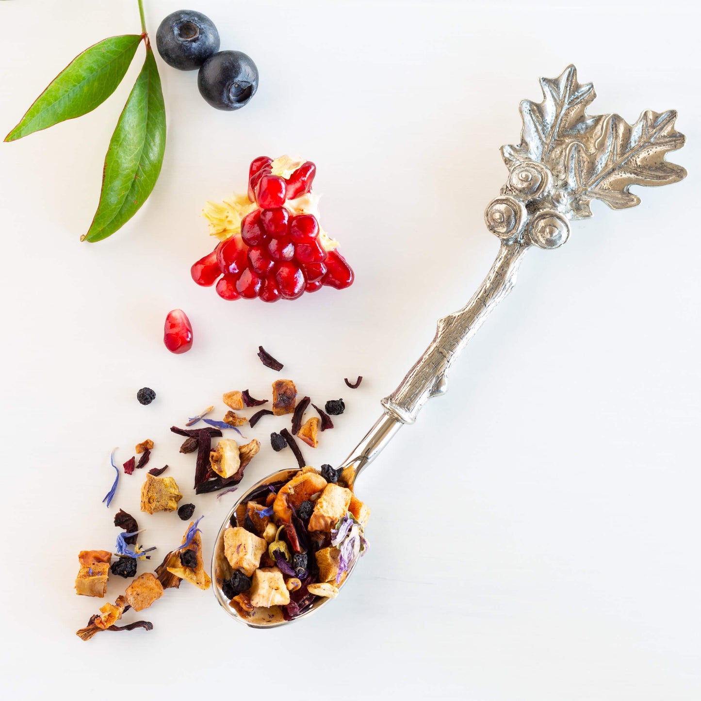 Blueberry Pomegranate Herbal Tea shown from above as loose tea leaves in a pewter spoon, with pomegranate arils and two blueberries nearby