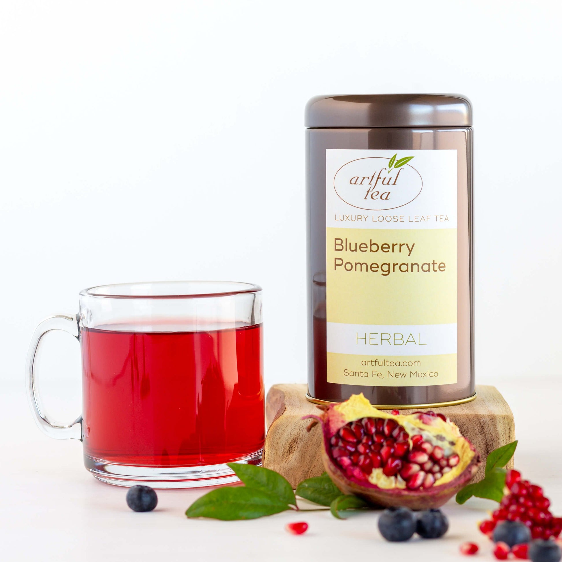 Blueberry Pomegranate Herbal Tea shown packaged in a brown tin, next to a glass mug of brewed tea, with an open pomegranate in the foreground 