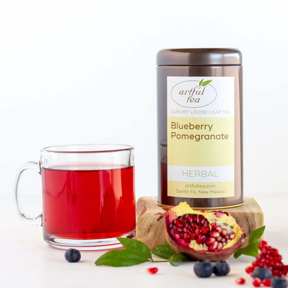 Blueberry Pomegranate Herbal Tea shown packaged in a brown tin, next to a glass mug of brewed tea, with an open pomegranate in the foreground 
