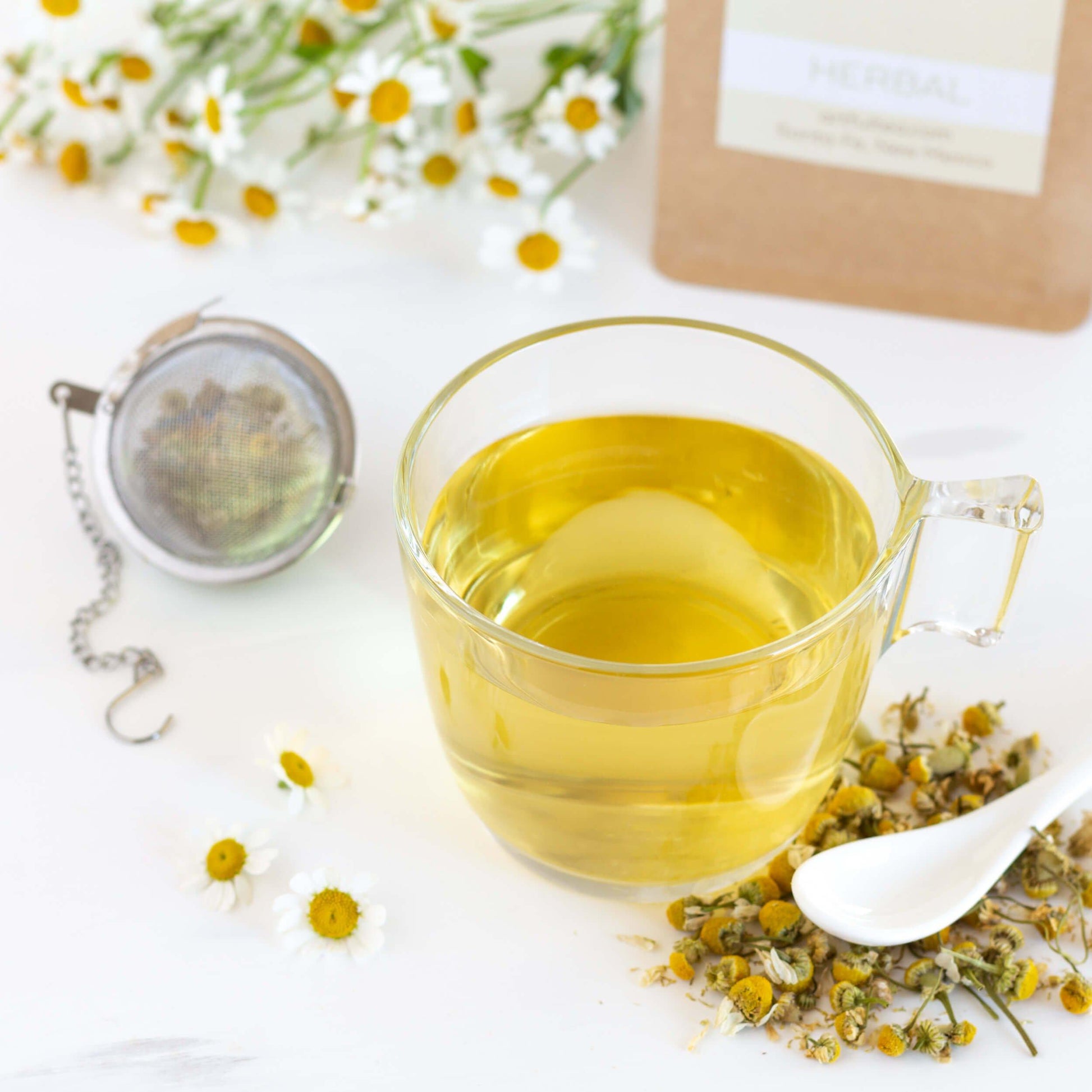 Egyptian Chamomile Organic Herbal Tea shown as brewed tea in a glass mug with a mesh teaball, loose chamomile flowers, and a white porcelain spoon